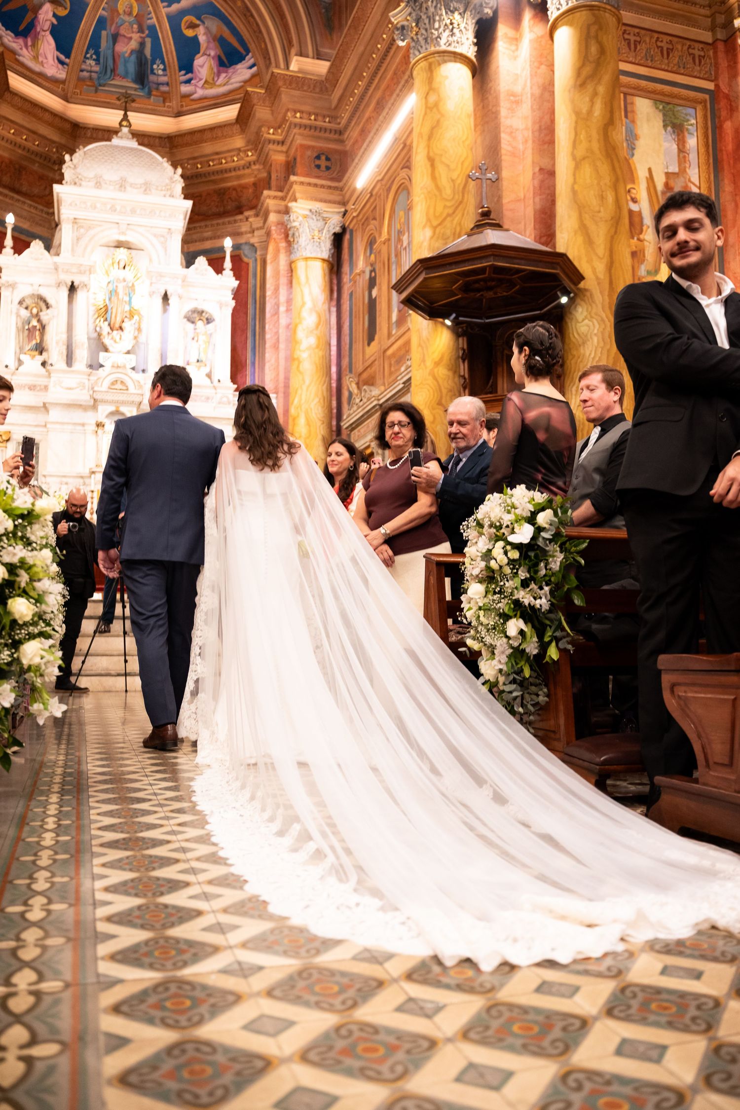 Bride in white gown with long veil walks down ornate church aisle with groom during wedding ceremony.