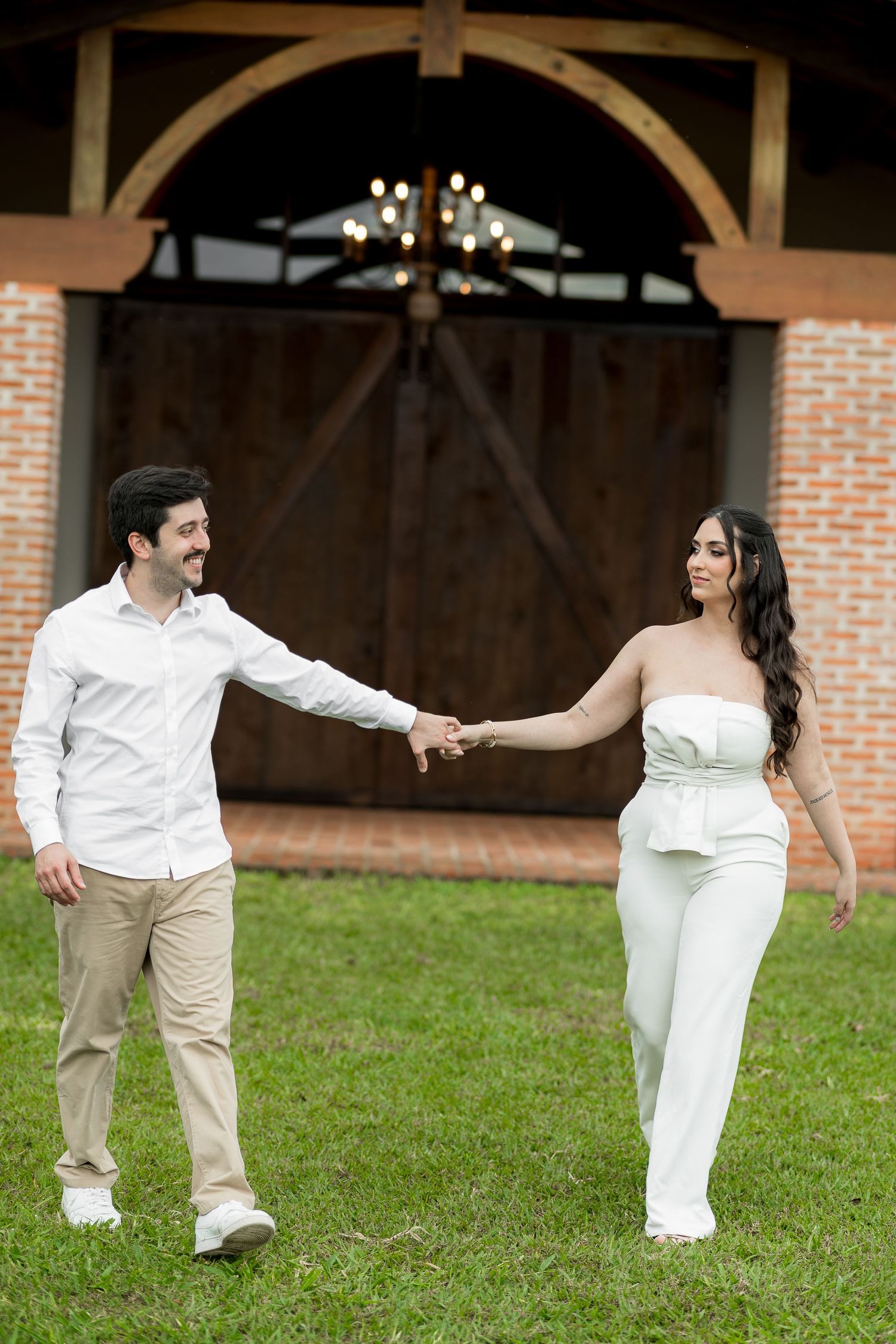 Couple holding hands on green grass near rustic barn doors, woman in white jumpsuit and man in white shirt and khaki pants.