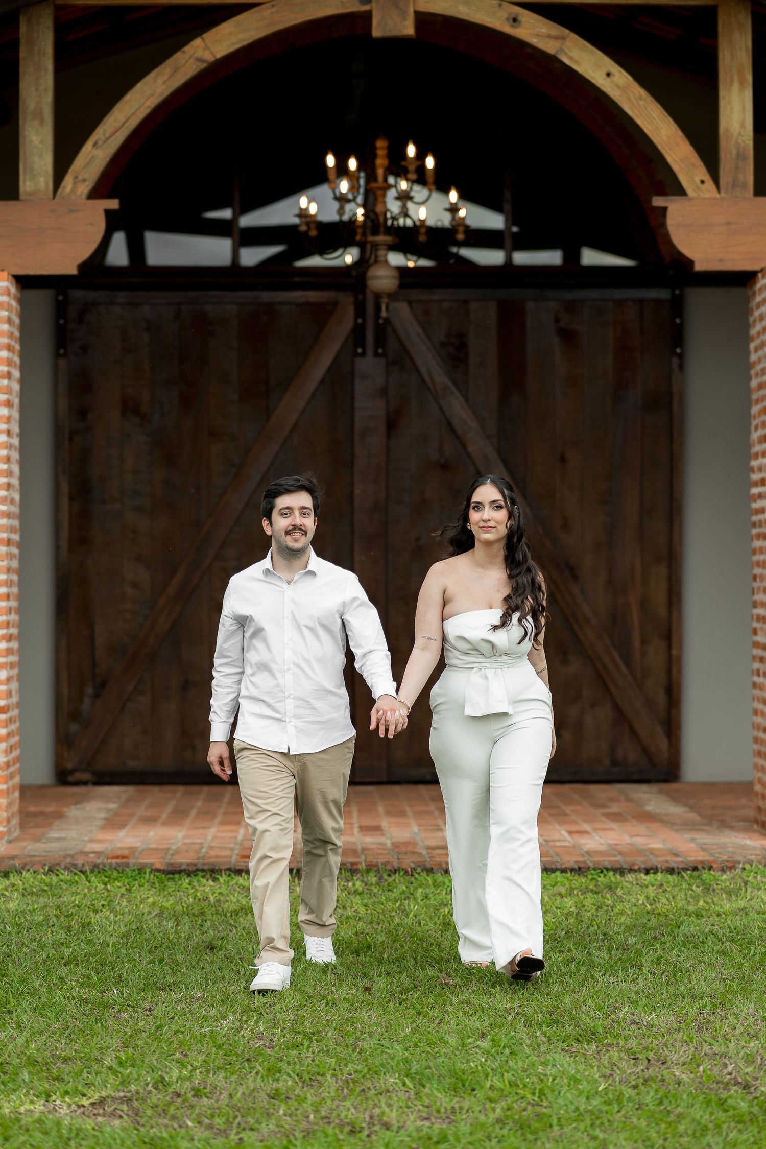 Couple holding hands walking on grass in front of rustic barn doors with chandelier, man in white shirt, woman in white jumpsuit.