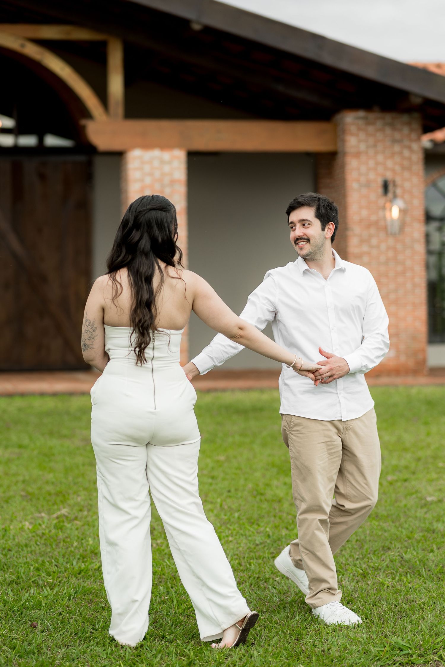 A couple in white outfits dance on green grass outside a brick building, smiling and holding hands.