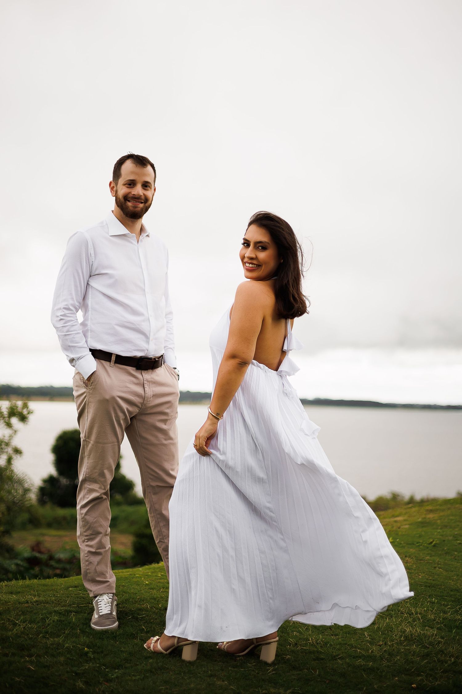 Smiling couple in white outfits posing outdoors near a lake, woman in flowing white dress, man in white shirt and khaki pants.