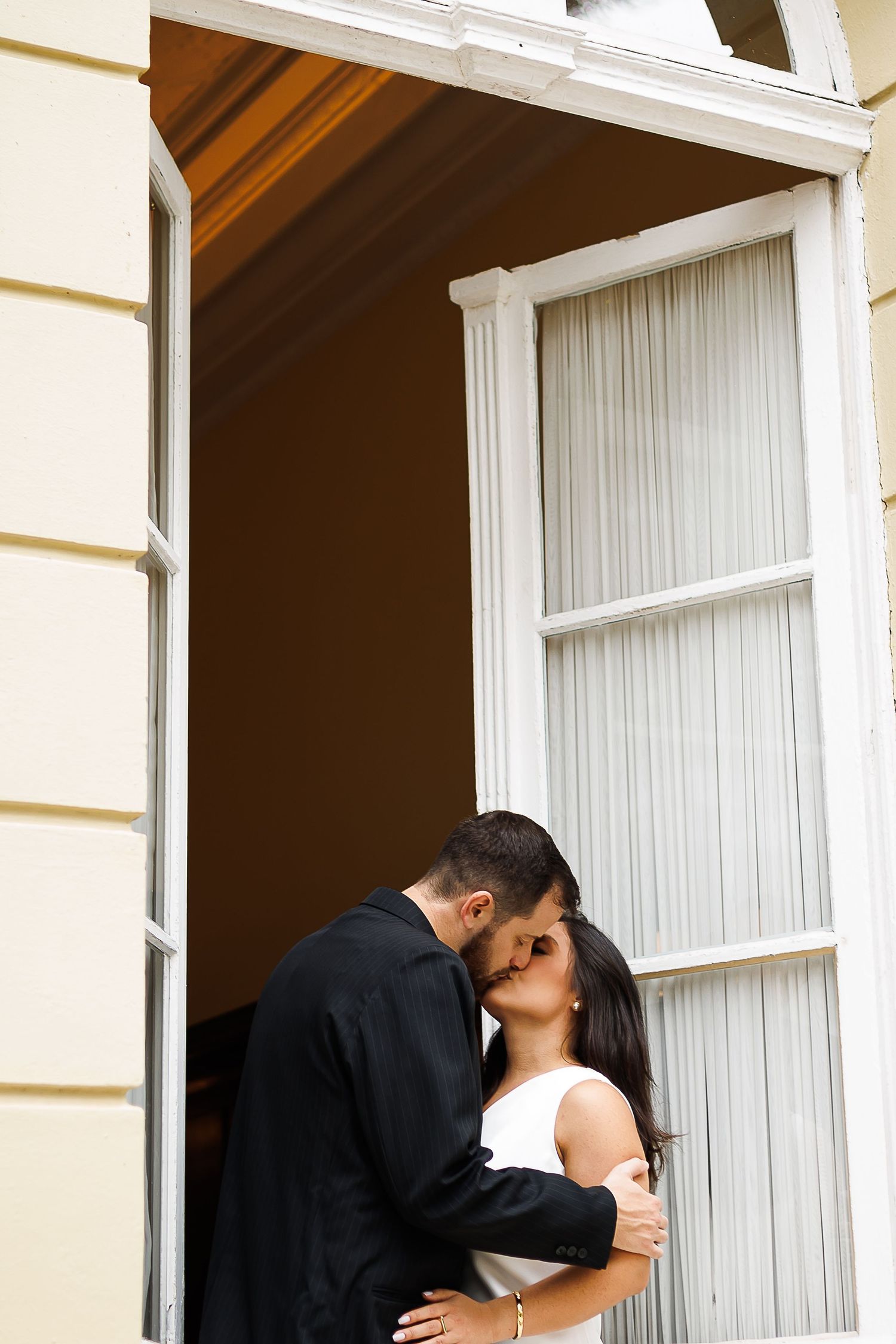 A couple shares a romantic kiss in a doorway, the groom in black and bride in white, framed by a yellow building.