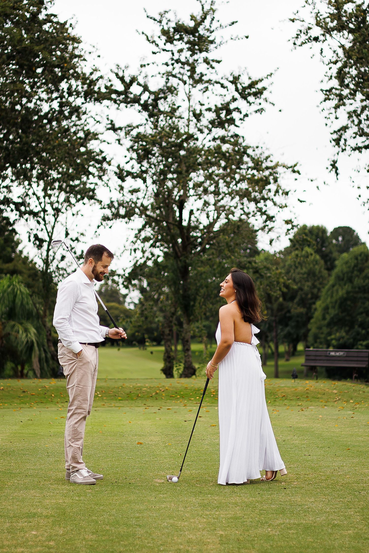Couple in white attire playing golf on a lush green course, sharing a joyful moment surrounded by trees.