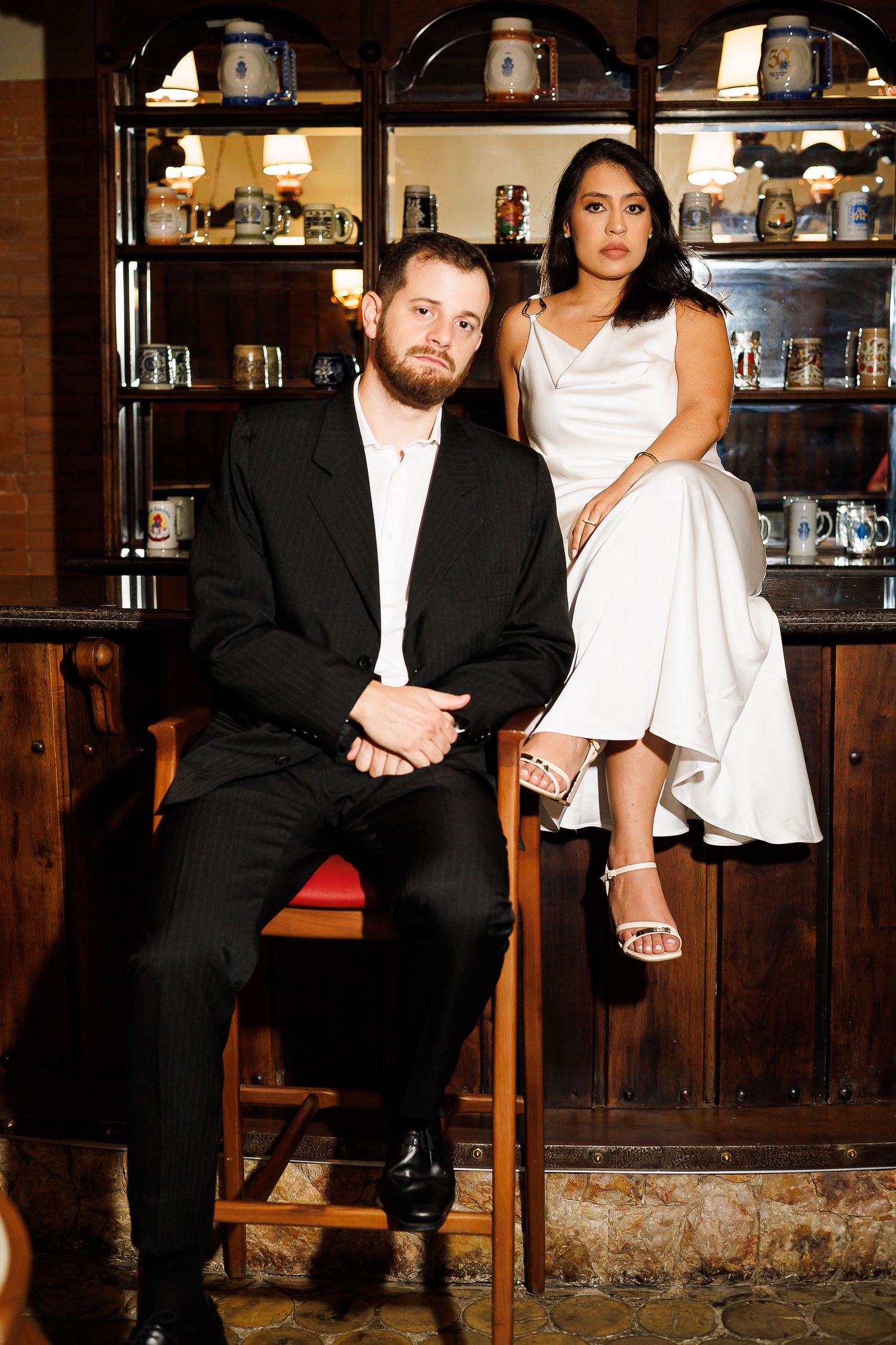 Couple in formal attire, man in black suit and woman in white dress, posing in a rustic bar setting.