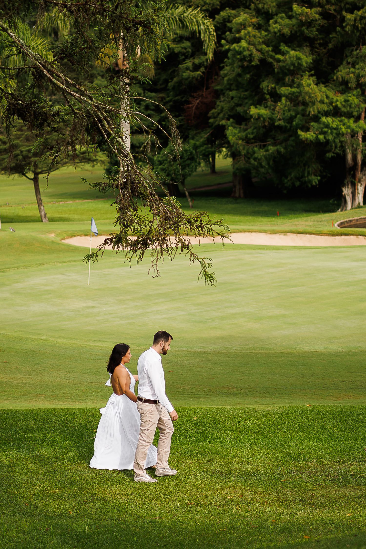 Couple in wedding attire walking hand in hand on a lush green golf course with sand bunker and flag in background.