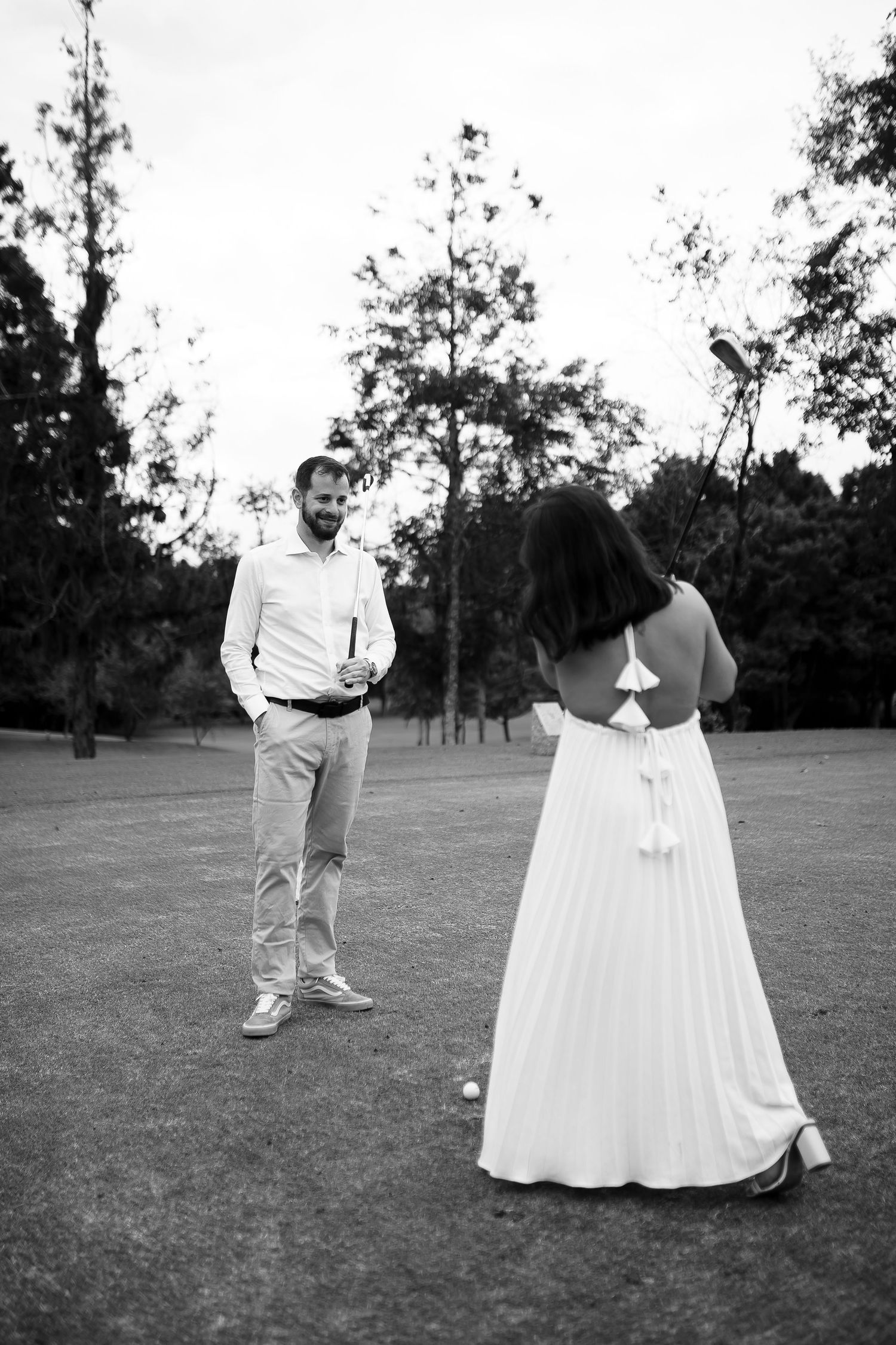 Black and white photo of a couple on a golf course, woman in white dress facing man in casual attire outdoors.