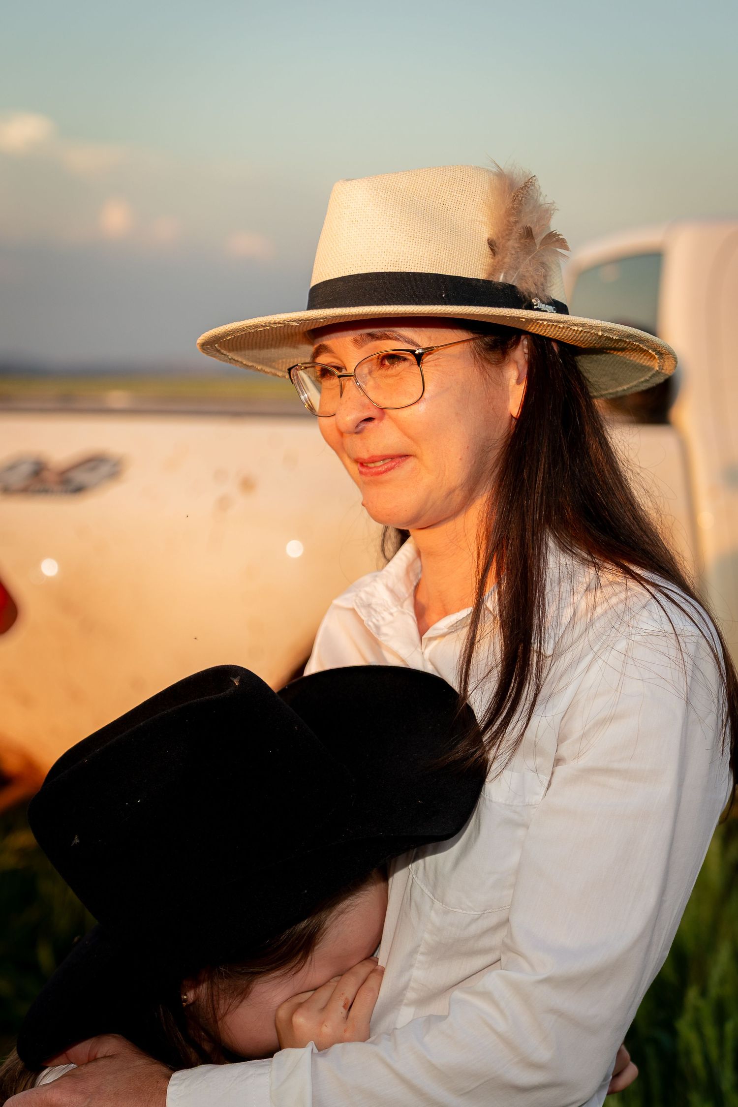 Woman in white shirt and straw hat with feather holds child outdoors at golden hour near a vehicle.