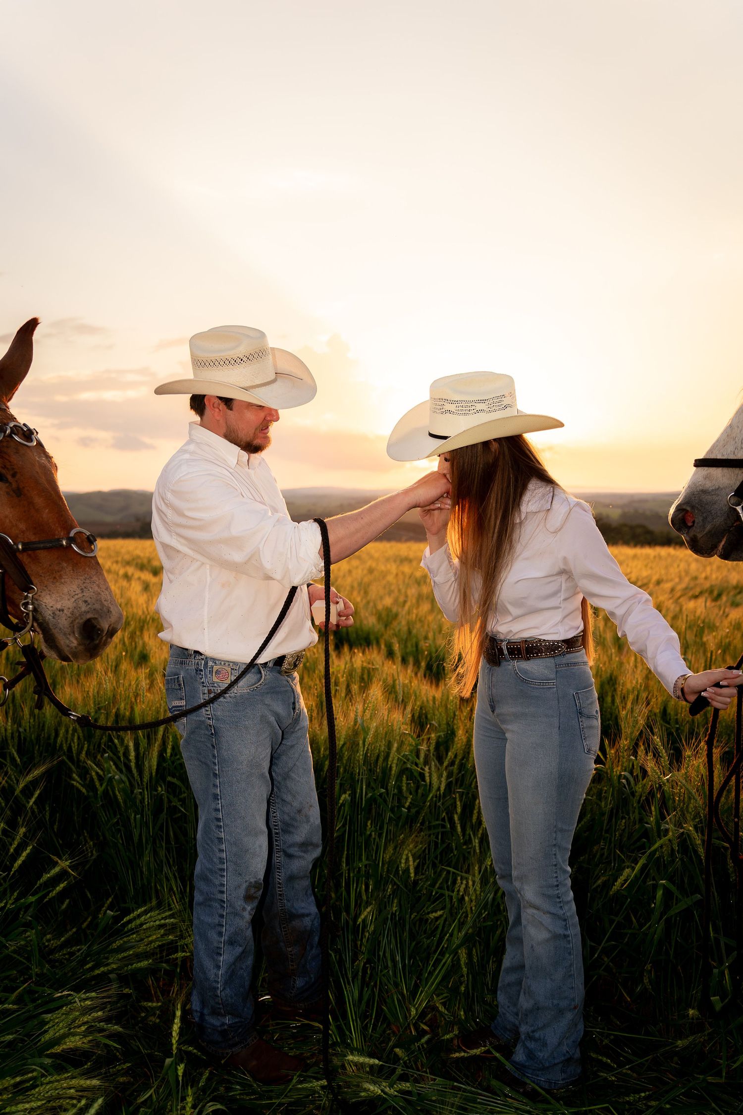Couple in cowboy hats and white shirts stand with horses in a golden wheat field at sunset.