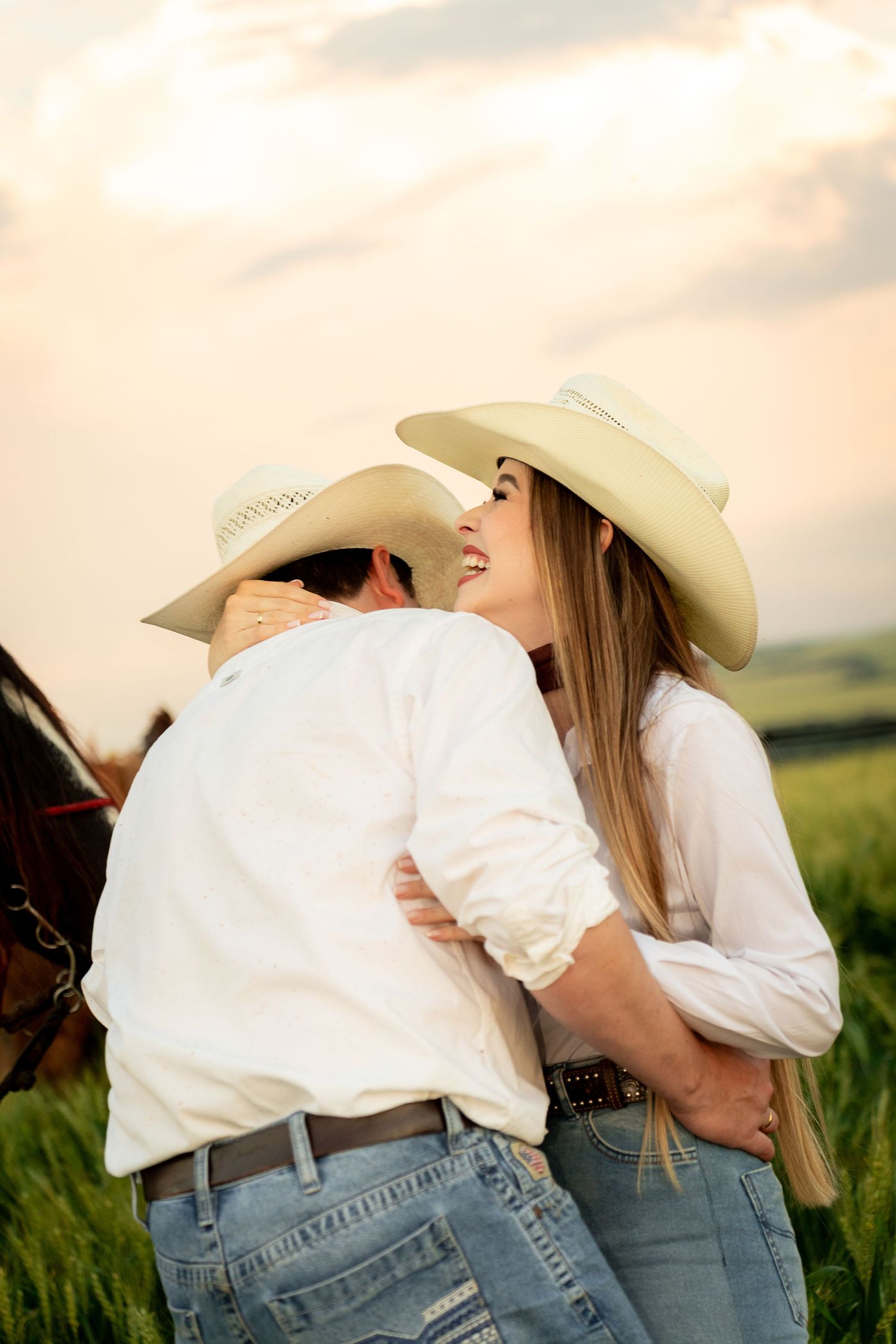 Couple in cowboy hats embracing in a green field at sunset, wearing white shirts and jeans near a horse.