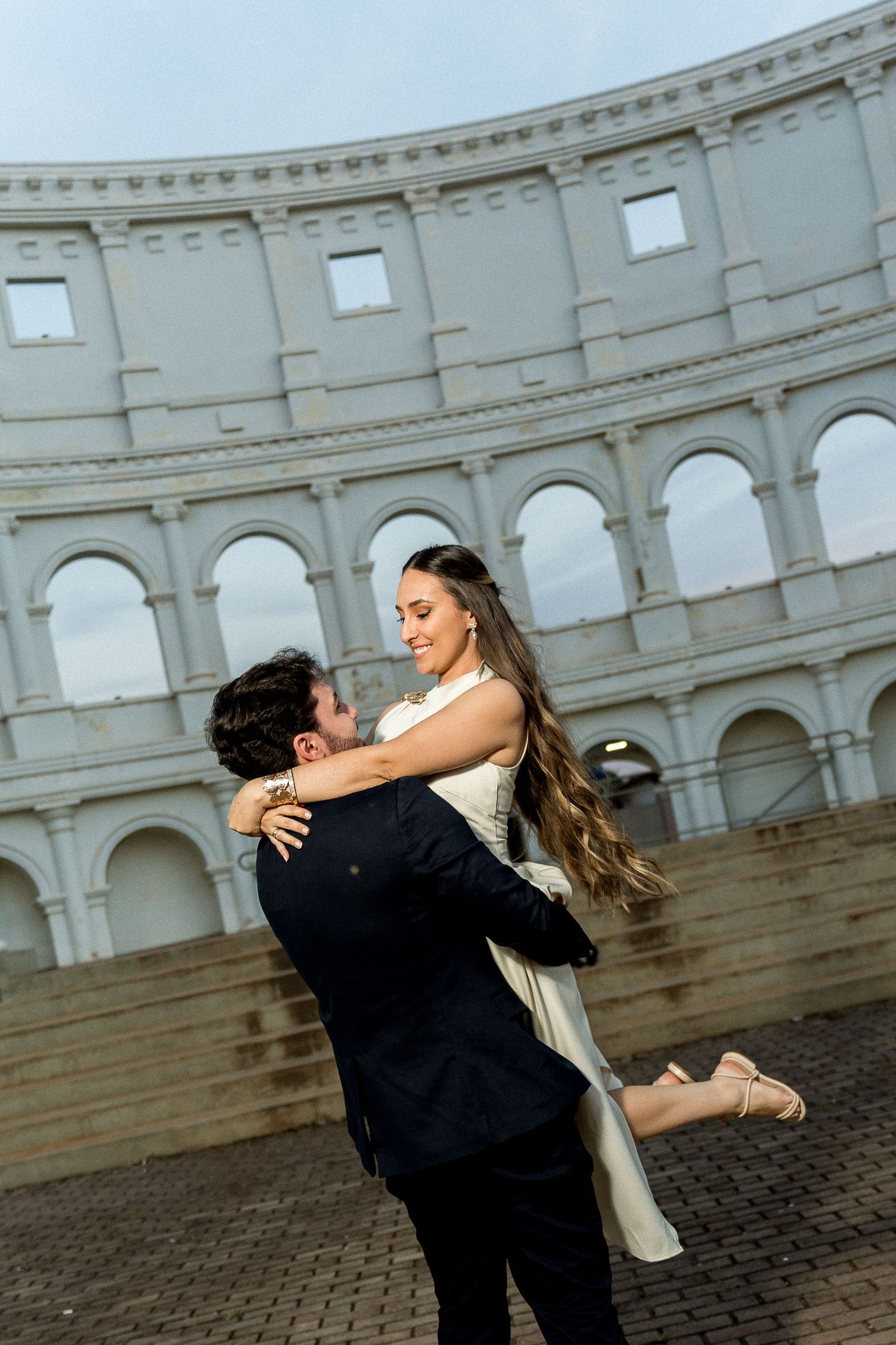 Couple embracing in front of the Colosseum in Rome, man lifting woman in white dress during romantic photo shoot.