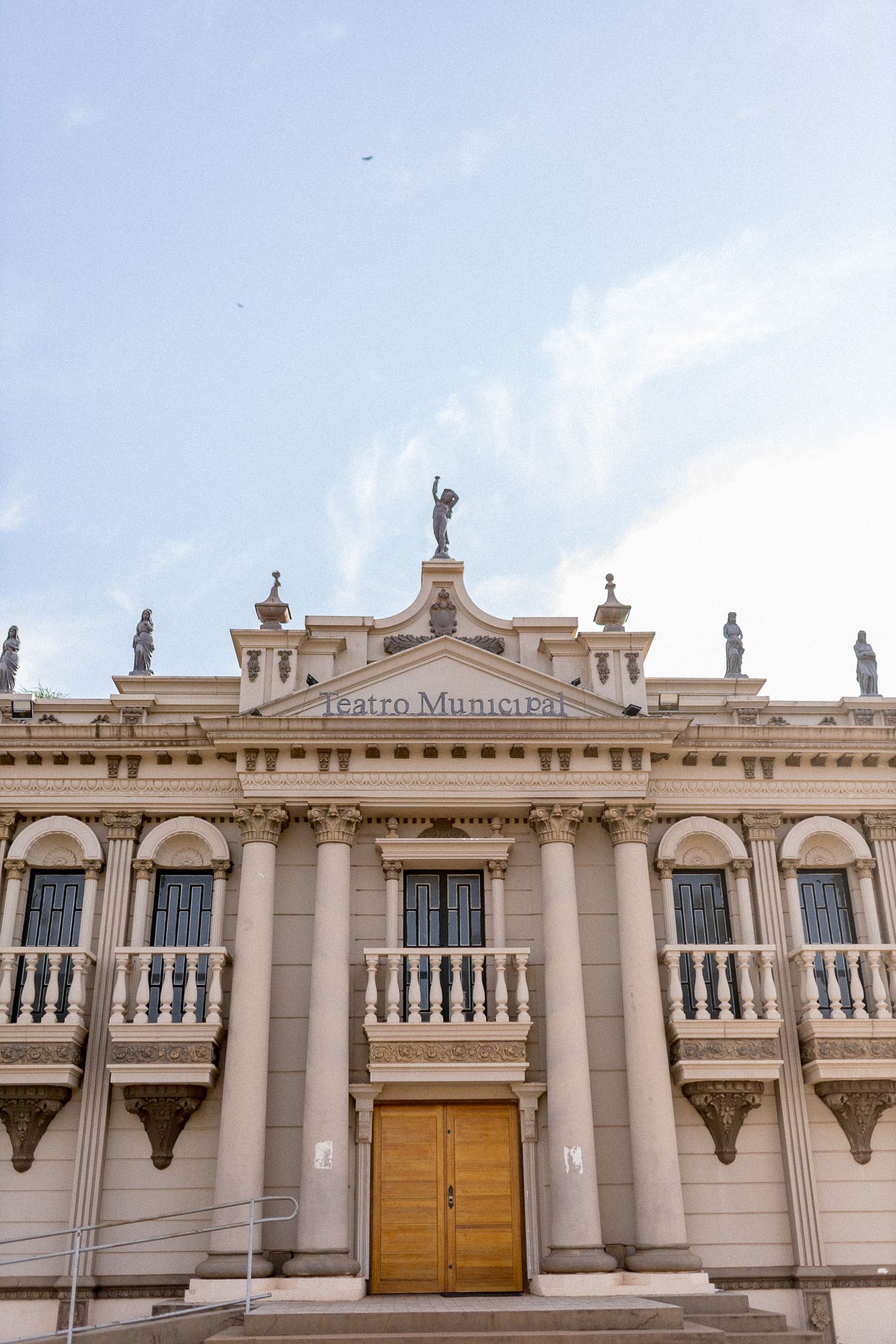 Neoclassical Teatro Municipal building facade with statues, columns, balconies, and wooden doors under a cloudy sky.