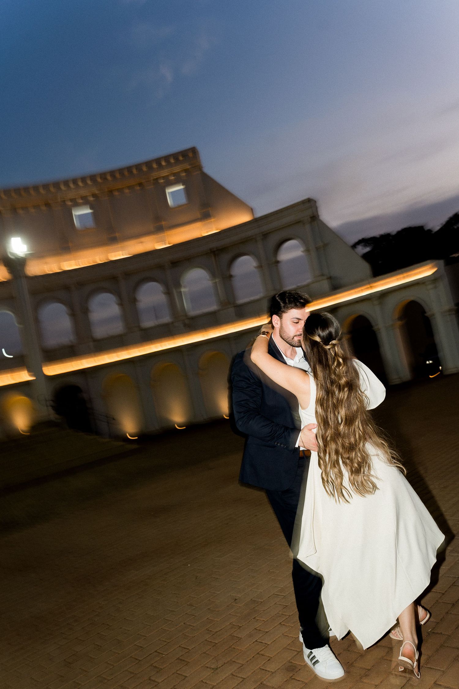 Couple dancing romantically outside the illuminated Colosseum in Rome at dusk.