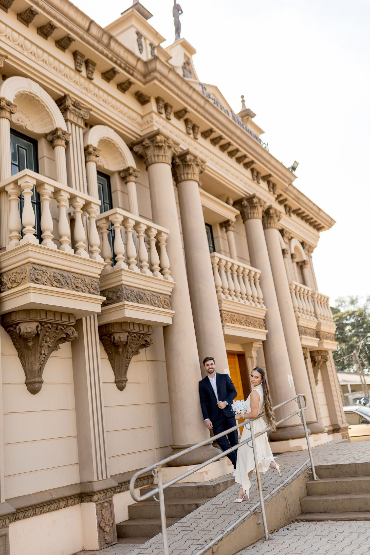 Bride and groom descend steps of elegant neoclassical building with columns and balustrades on their wedding day.