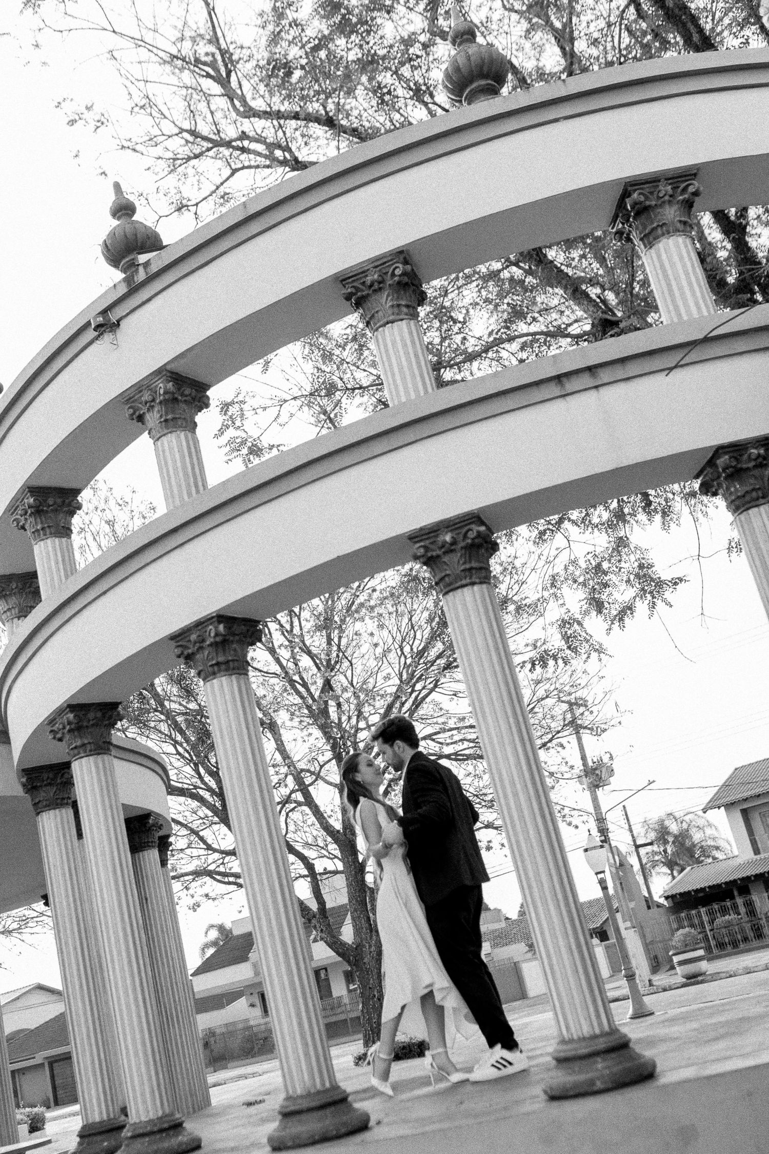 Black and white photo of a couple sharing a romantic moment under a elegant circular columned rotunda outdoors.