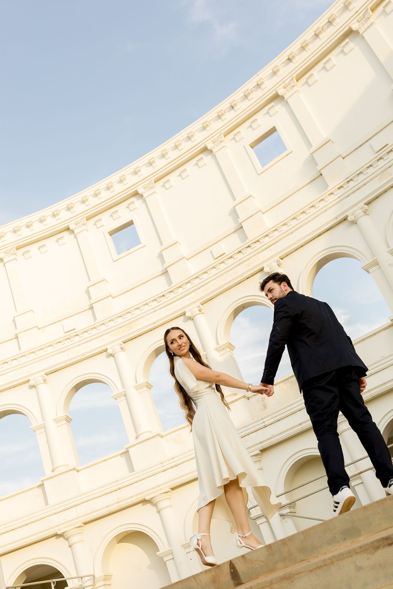 Couple holding hands in front of the Colosseum, woman in white dress and man in black suit.