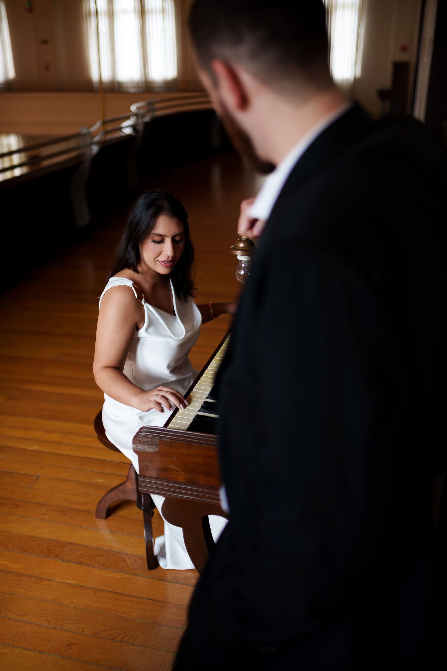 Woman in white dress playing piano while man in black suit watches in elegant wood-floored hall.