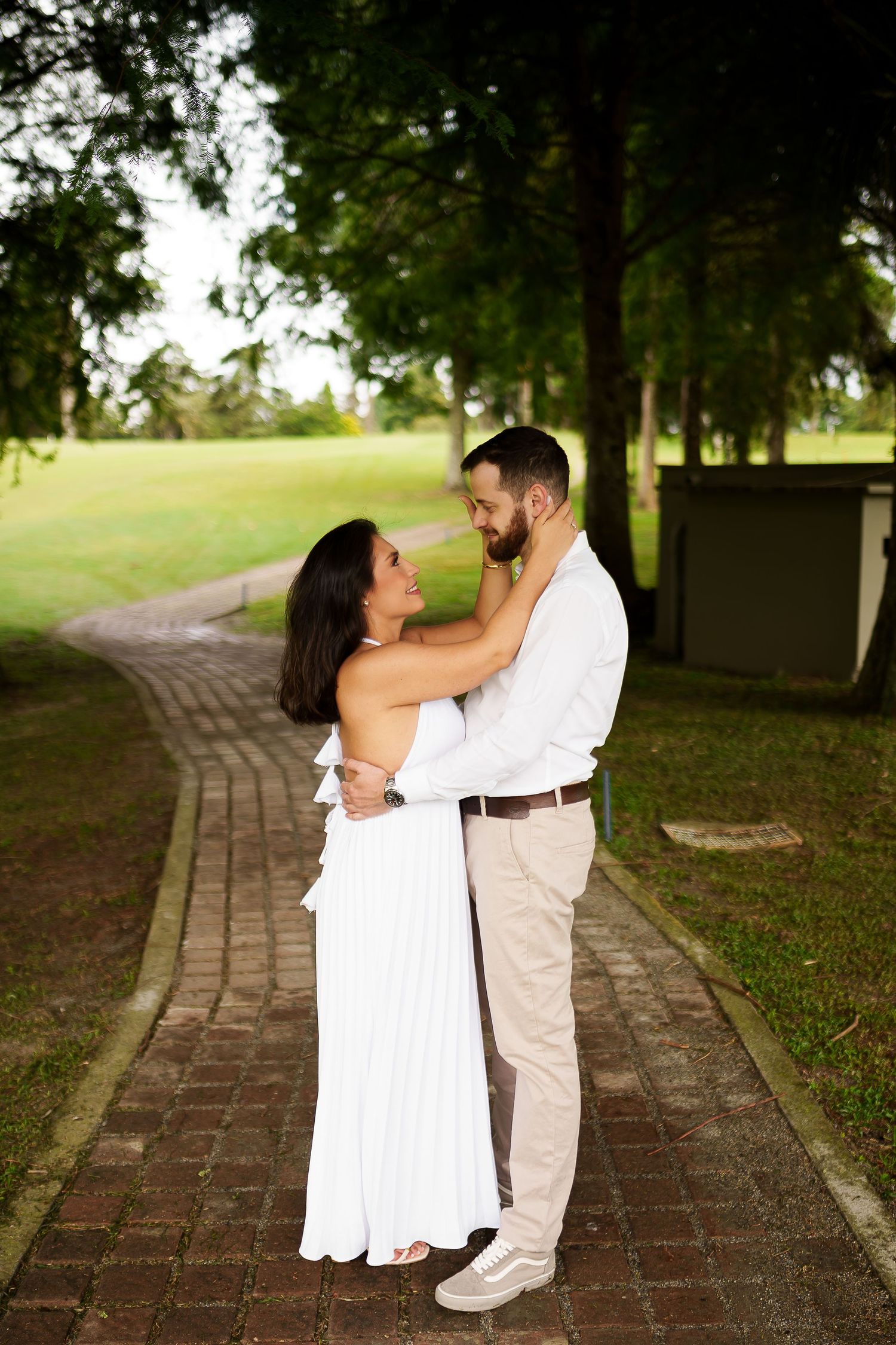 Couple in white outfits embracing on a brick path in a lush green park setting.