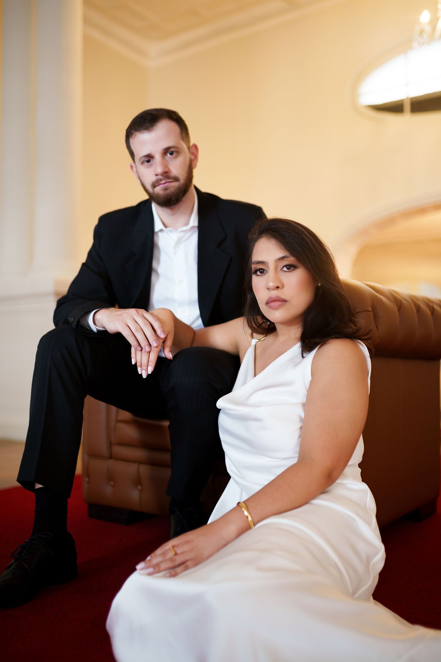 Elegant couple in formal attire, man in black suit and woman in white dress, posing in a luxurious room.