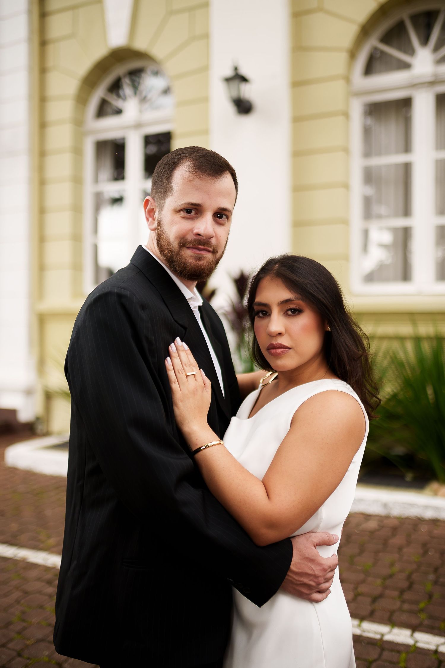 Elegant couple in formal attire embracing outside a yellow building, woman in white dress, man in black suit.