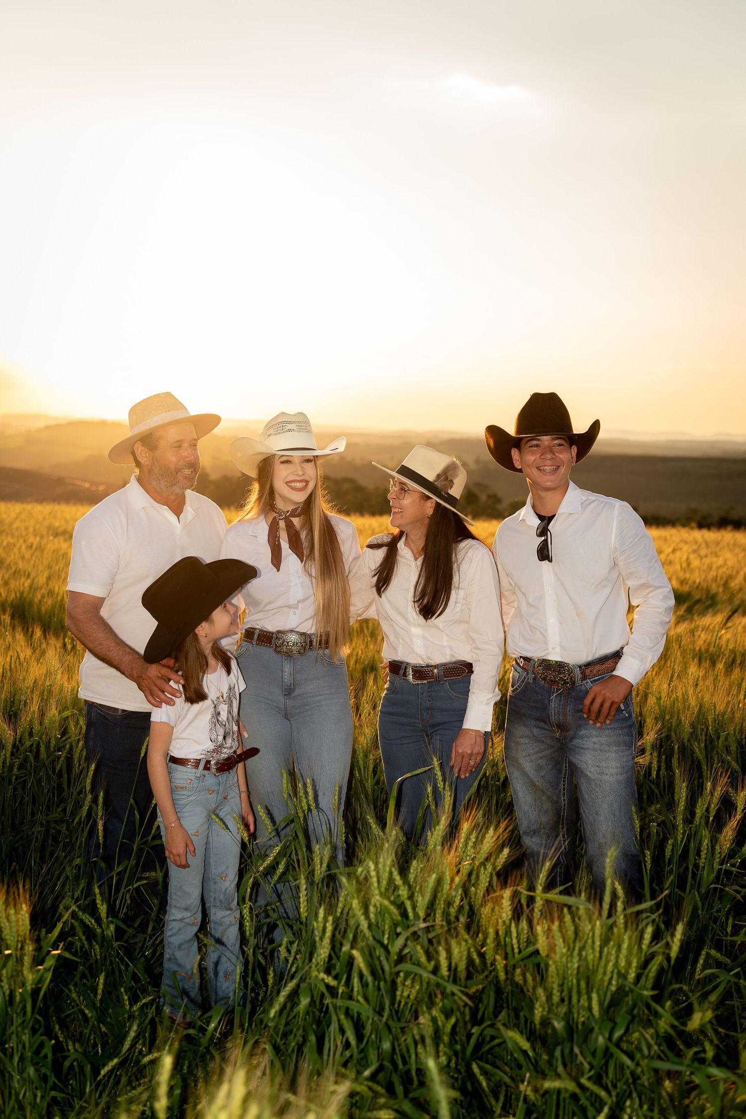 Family of five in cowboy hats and white shirts posing in a golden wheat field at sunset.