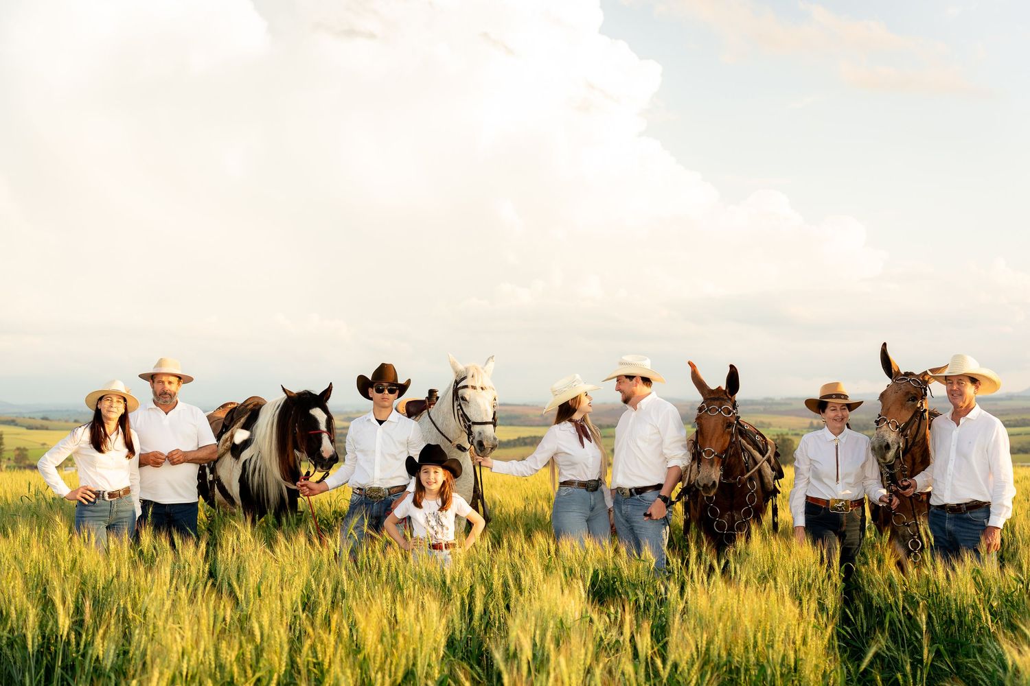 A group of cowboys and cowgirls in straw hats pose with horses in a golden wheat field under a cloudy sky.
