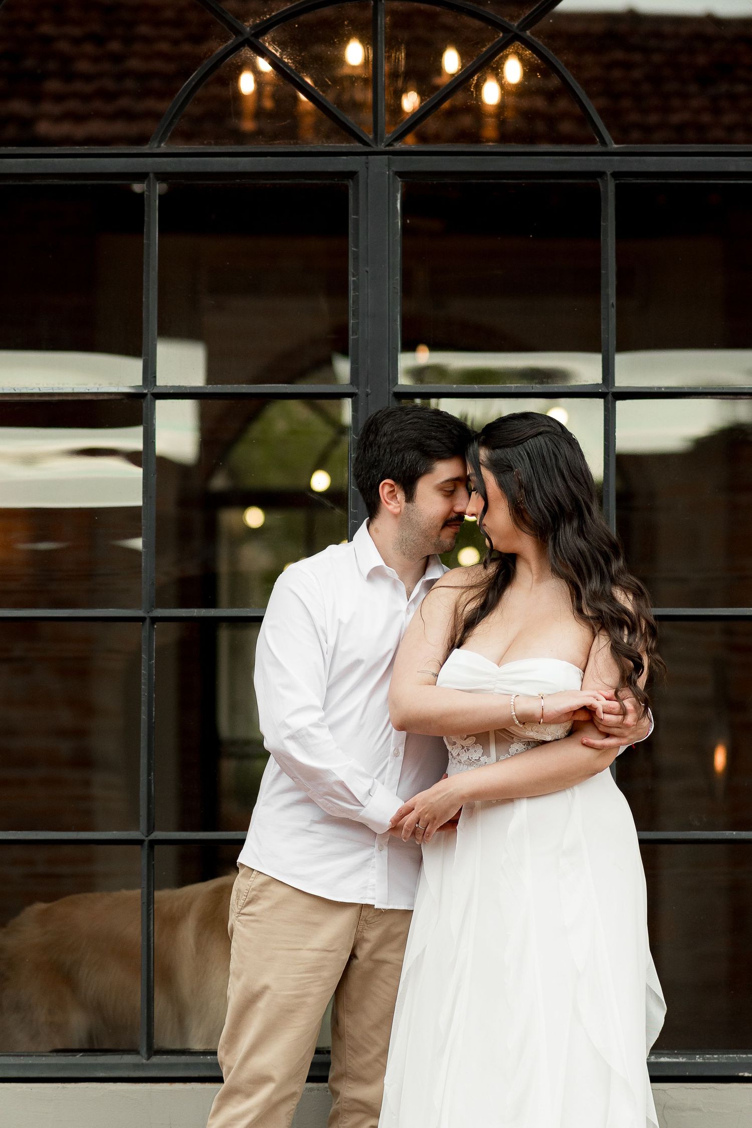 Romantic couple in white dress and khaki pants embracing before arched black window with warm string lights.