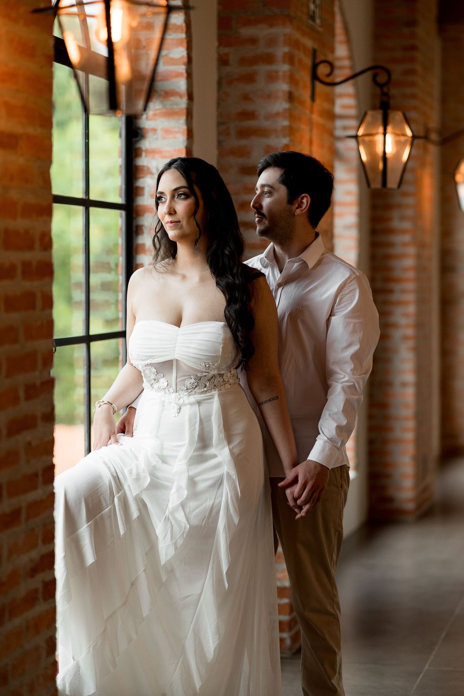 Elegant bride in white strapless gown holds hands with groom in white shirt on brick corridor with lanterns.