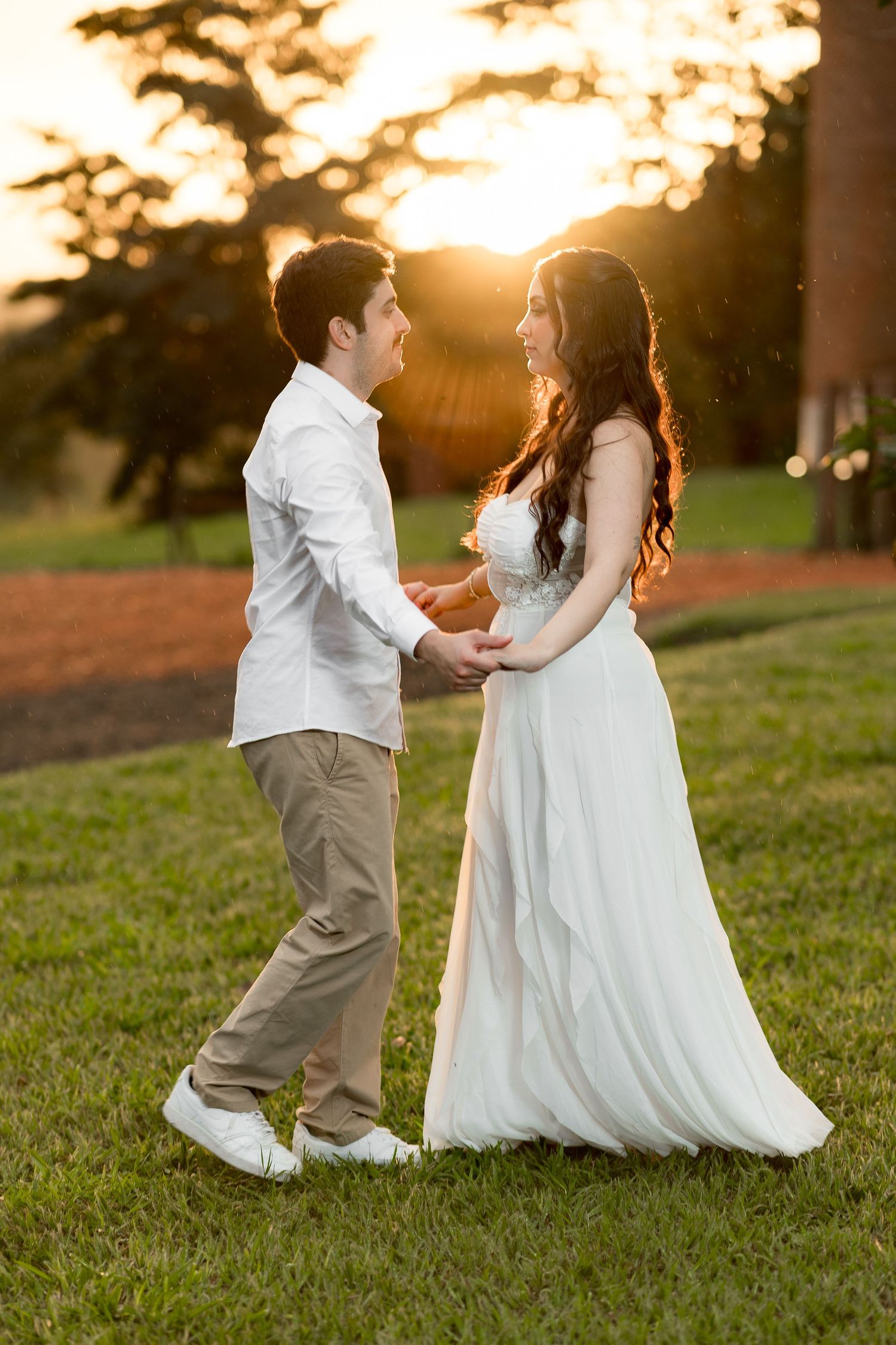 Couple in white wedding attire holding hands at golden sunset in lush green park.