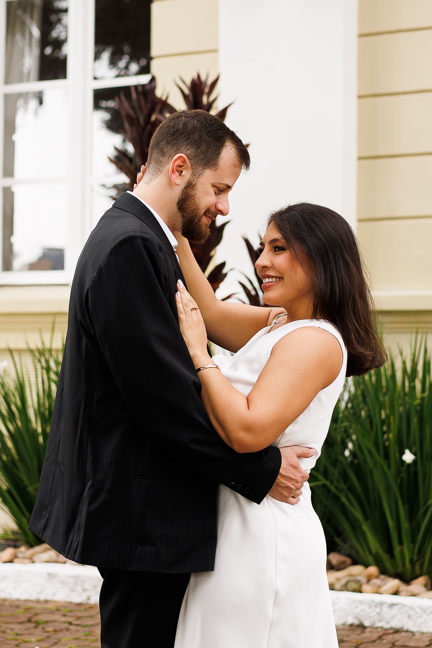A smiling couple in wedding attire embrace outdoors, with the bride in white and groom in a black suit.