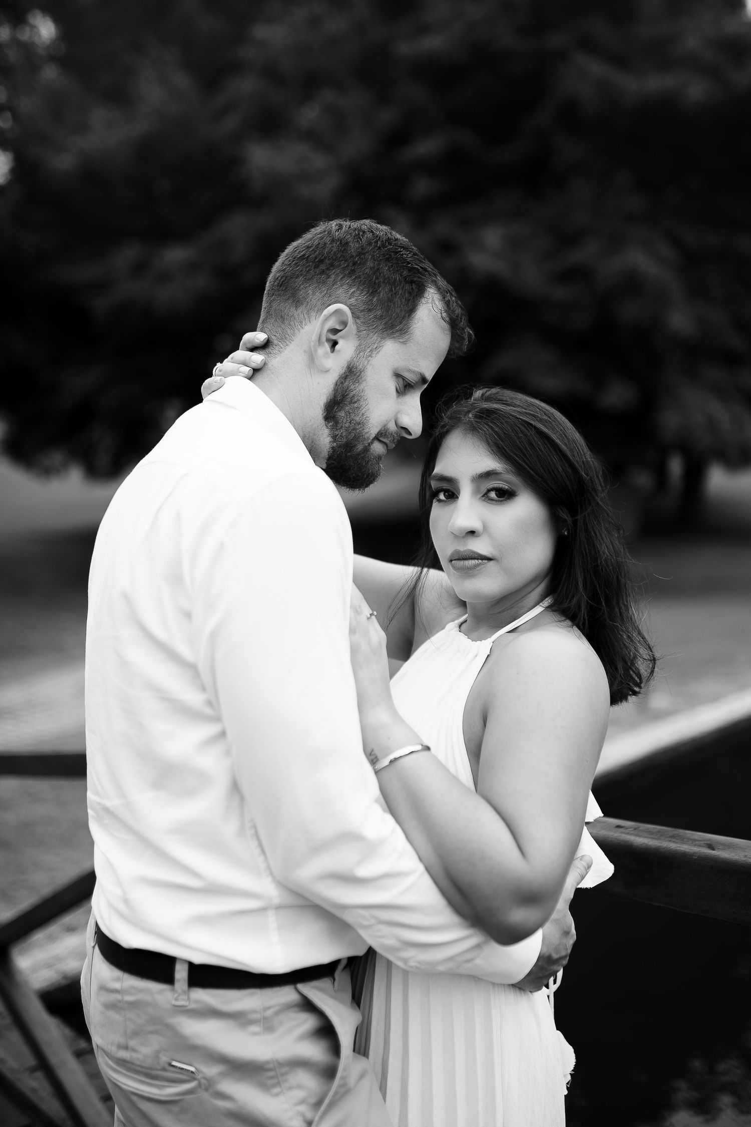 Black and white photo of a couple embracing outdoors, woman in white dress looking at camera, man in white shirt.