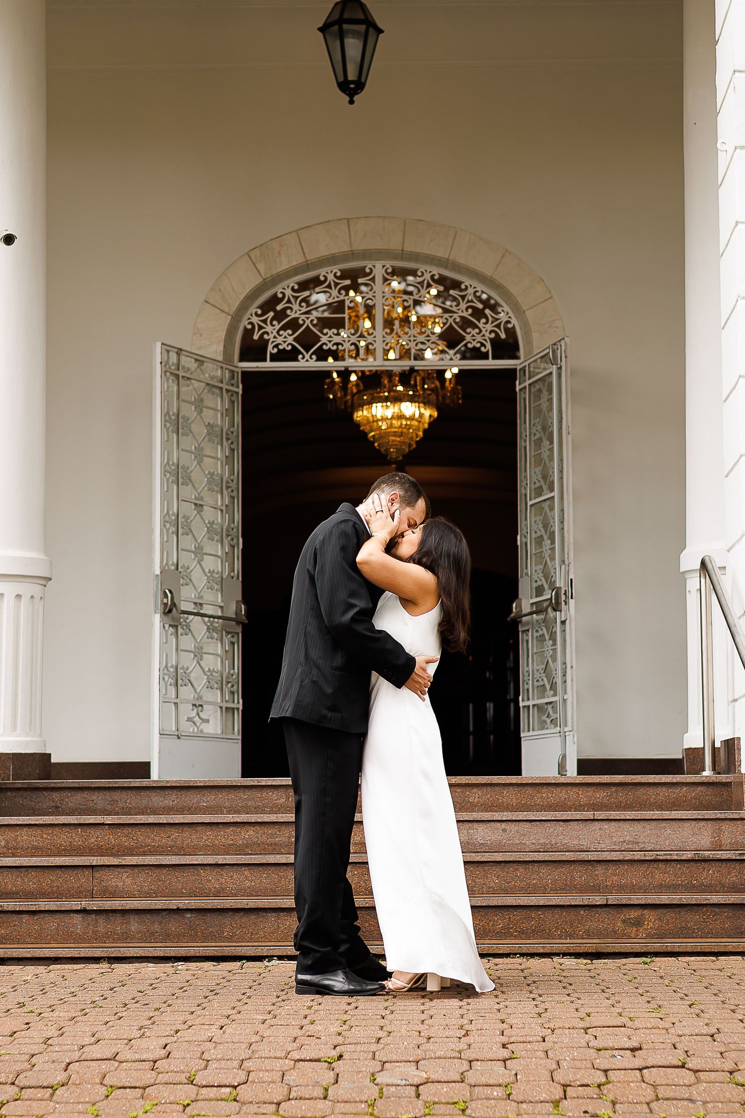Bride and groom share a romantic kiss on steps outside elegant white building with ornate chandelier and glass doors.