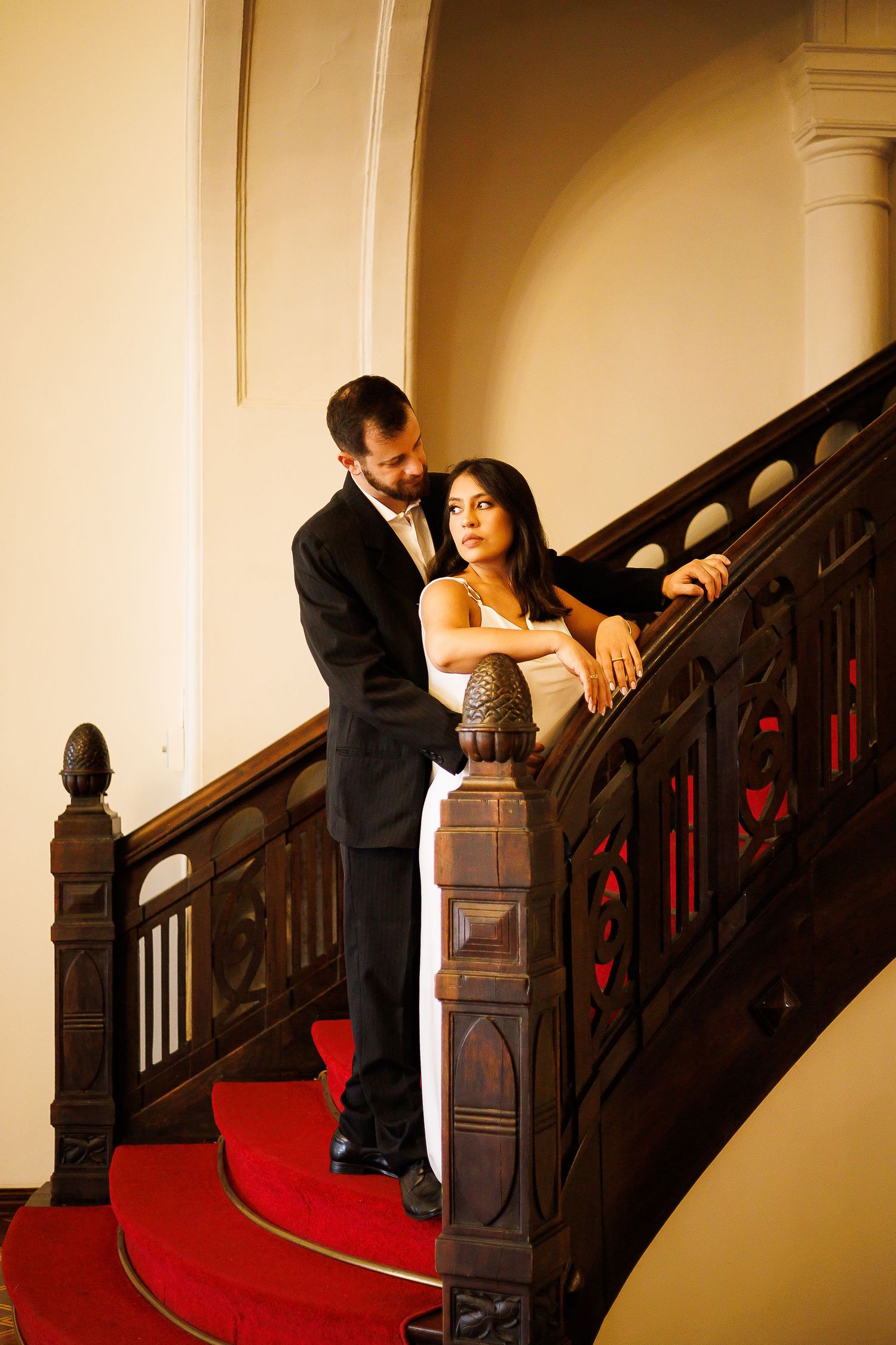 Elegant couple posing on ornate wooden staircase with red carpet in a grand interior setting.