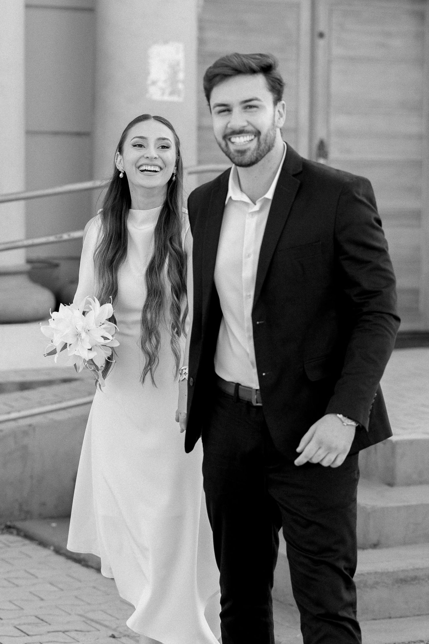 Black and white photo of a smiling bride in white dress holding bouquet standing with groom in dark suit on steps.