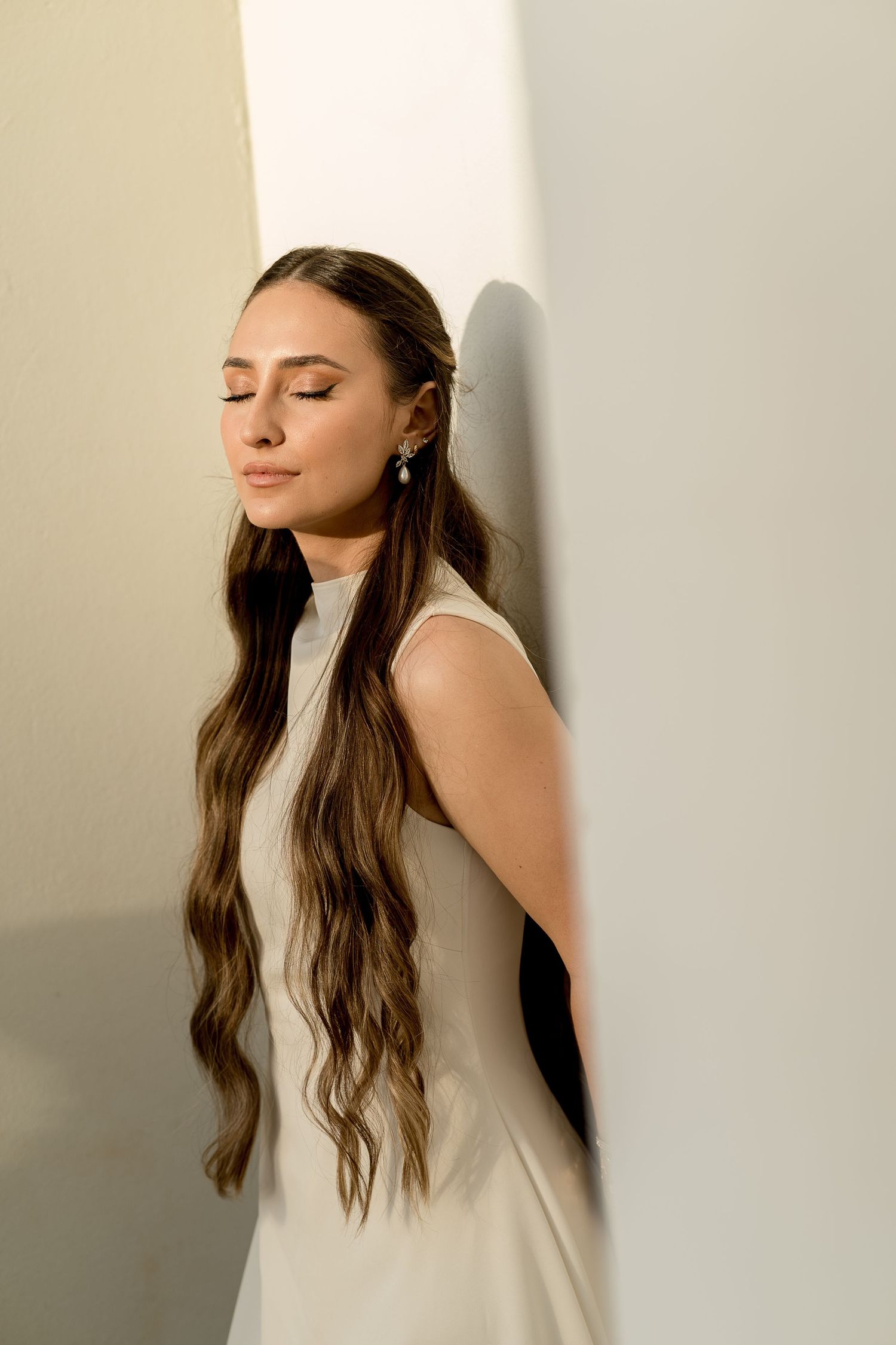 Young woman with long wavy hair in white dress leans against wall, eyes closed, wearing pearl earring.