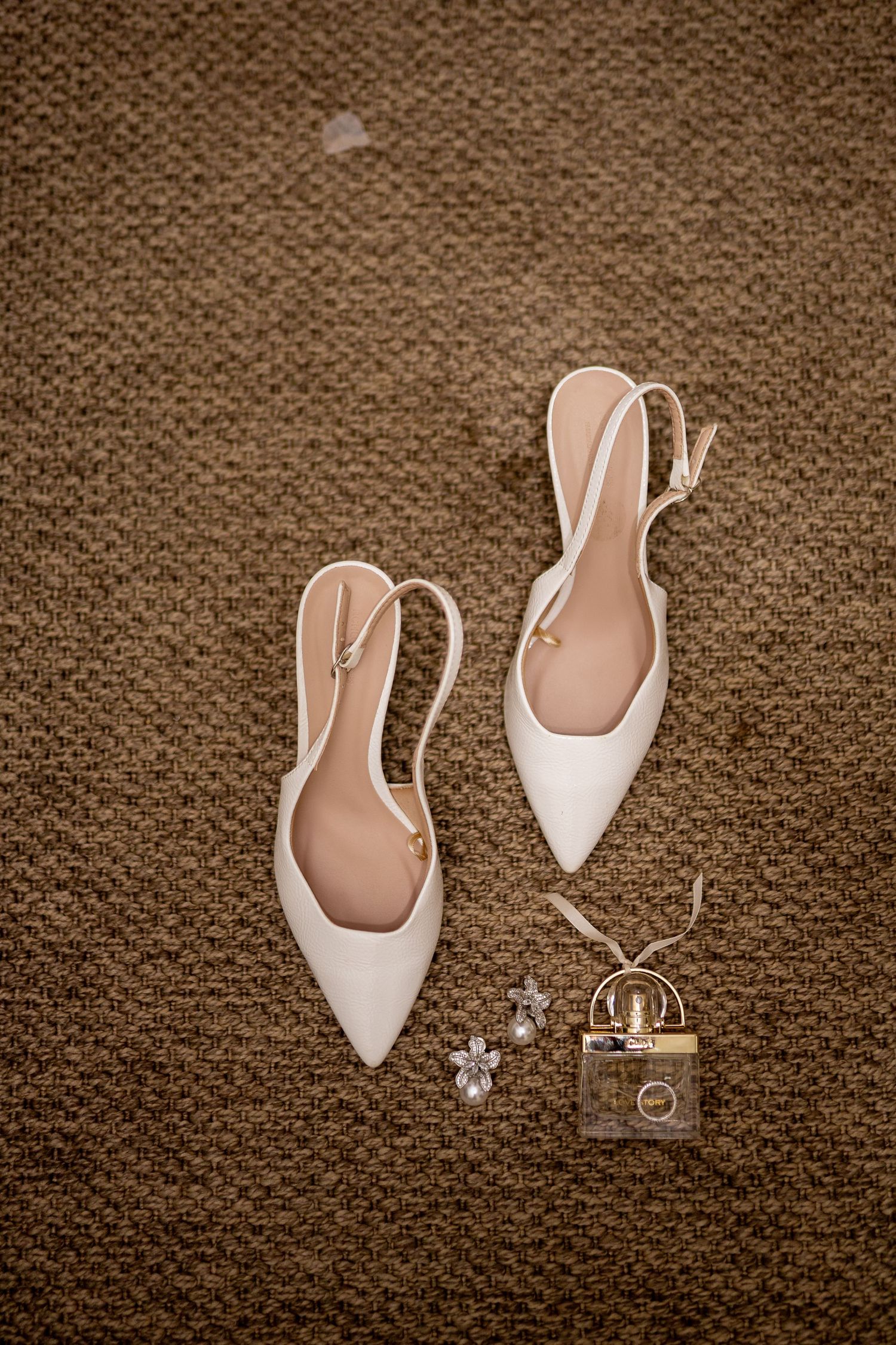 White slingback pointed-toe bridal heels displayed on carpet beside pearl earrings and gold padlock charm accessory.