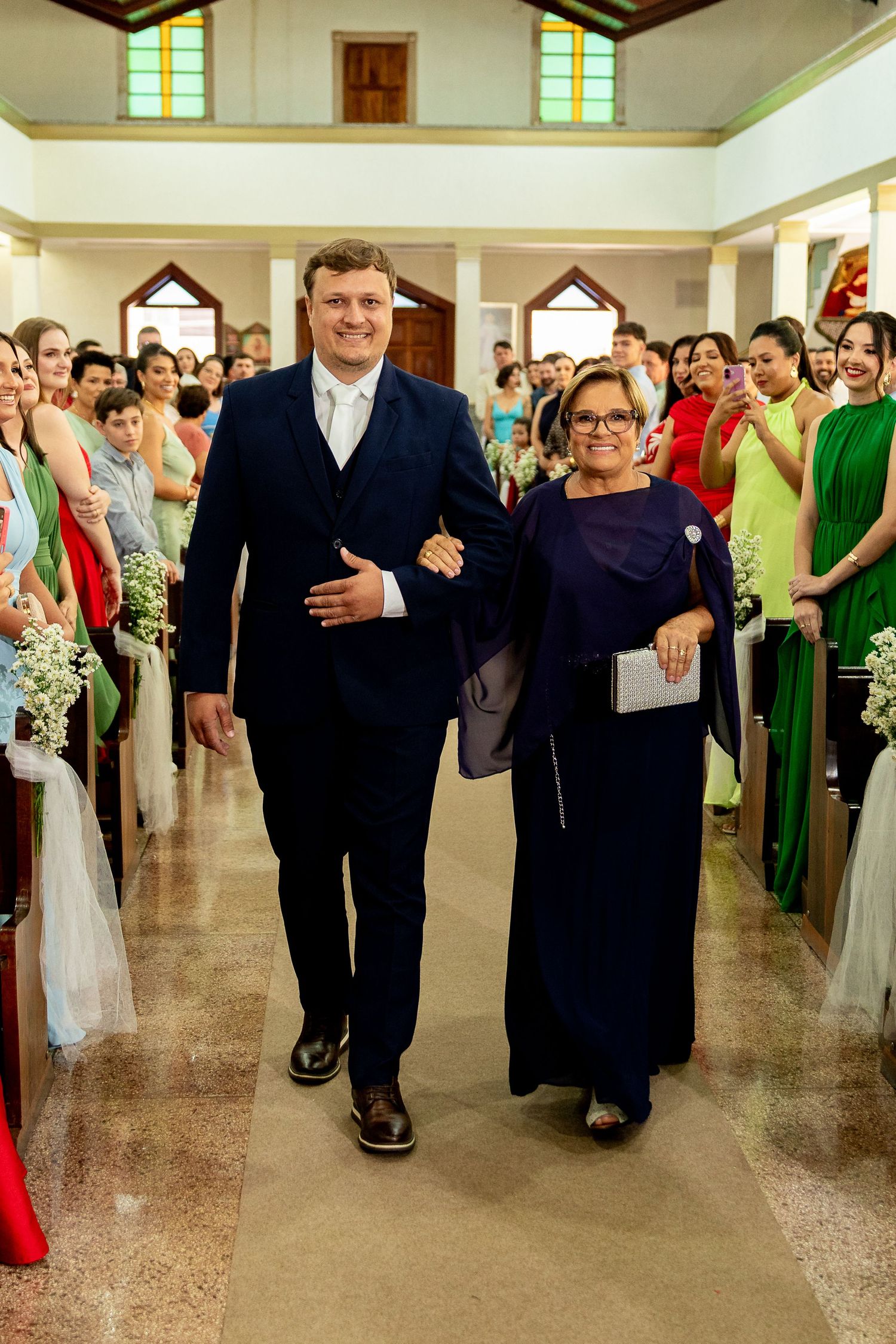 Groom in navy suit walks down church aisle with older woman in blue dress during wedding ceremony.