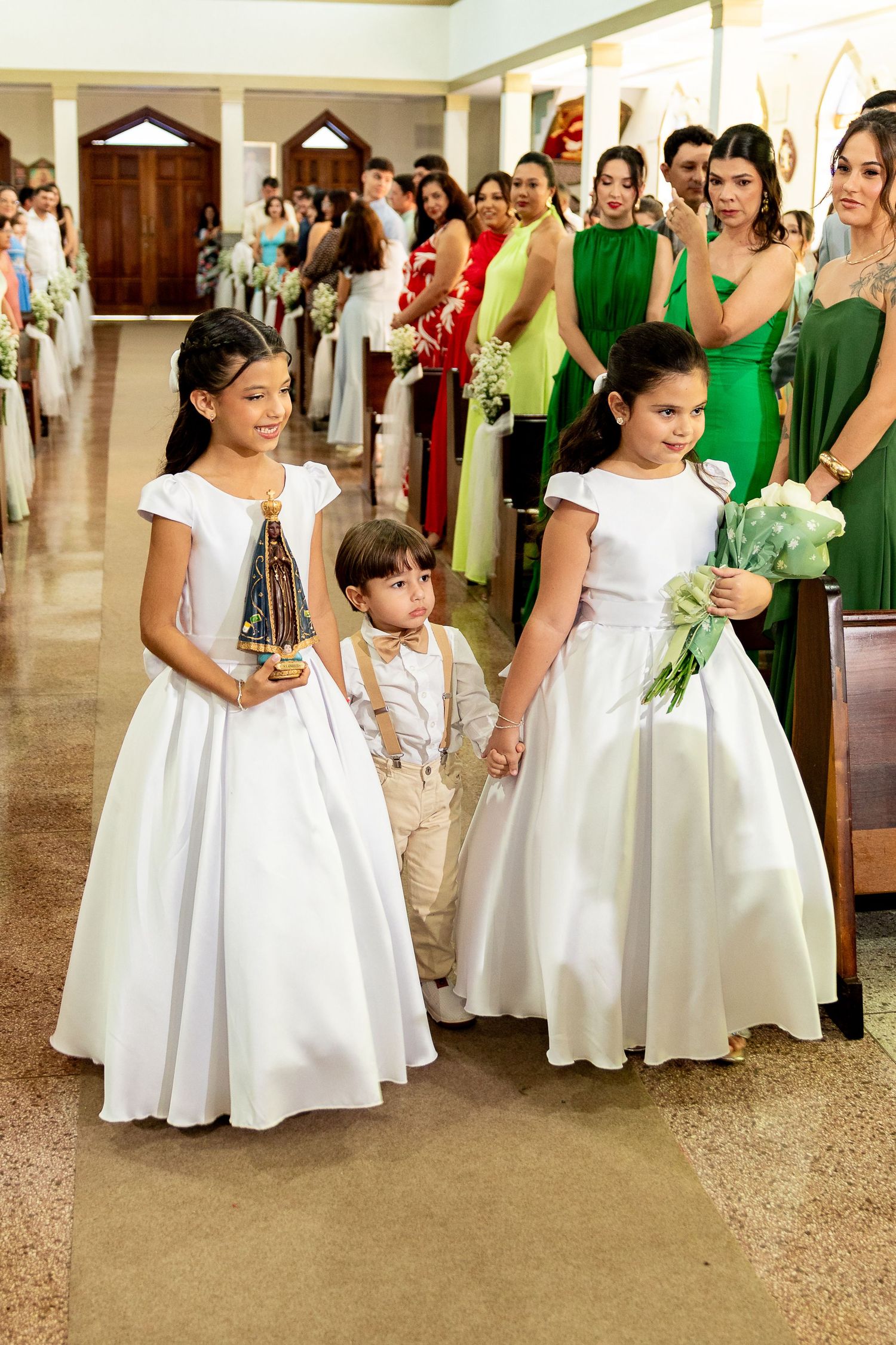 Two flower girls in white dresses and a ring bearer walk down a church aisle during a wedding ceremony.