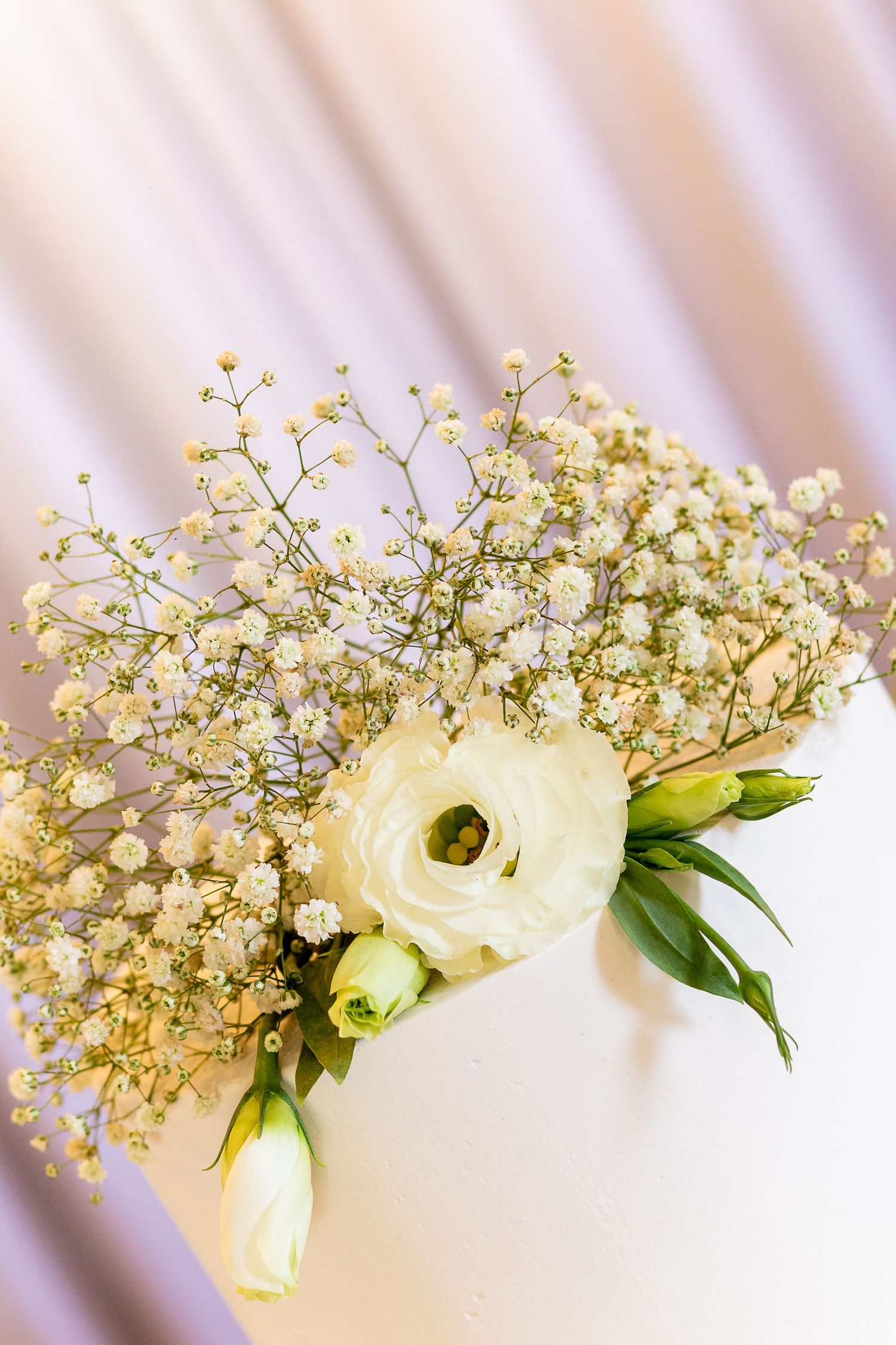 White ranunculus and baby's breath floral arrangement on a white cake with soft pink draped fabric background.