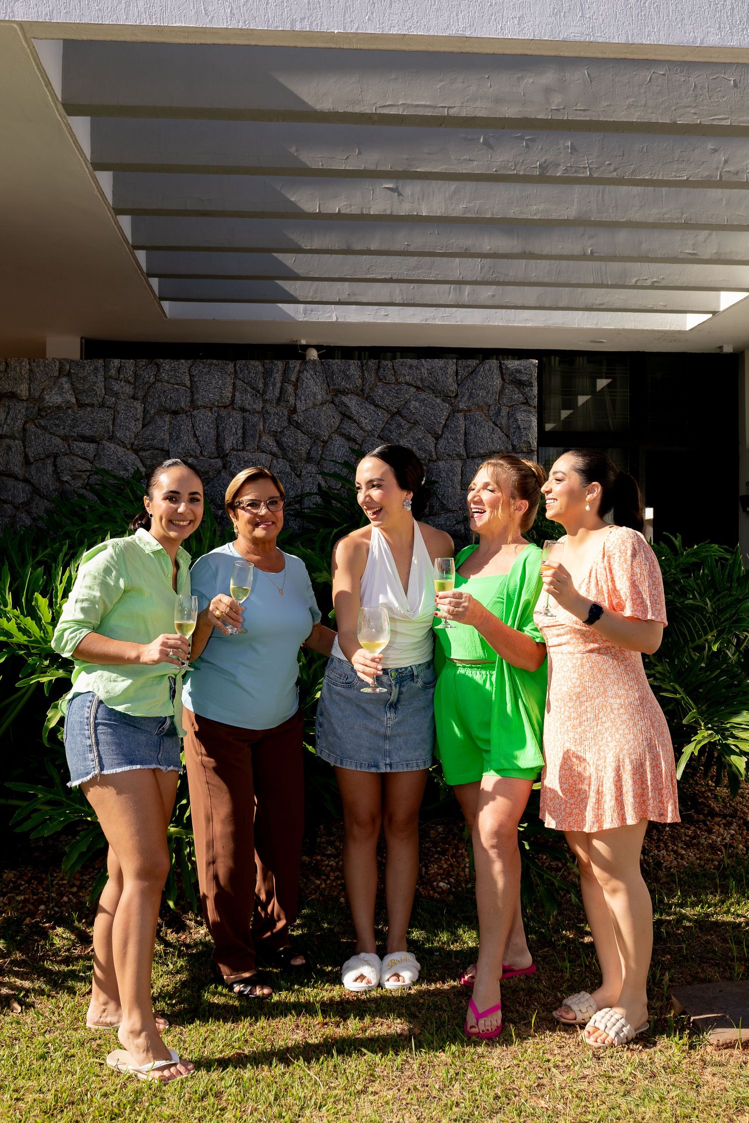 Five women smiling and holding champagne glasses outdoors in colorful summer outfits, celebrating together in a garden.