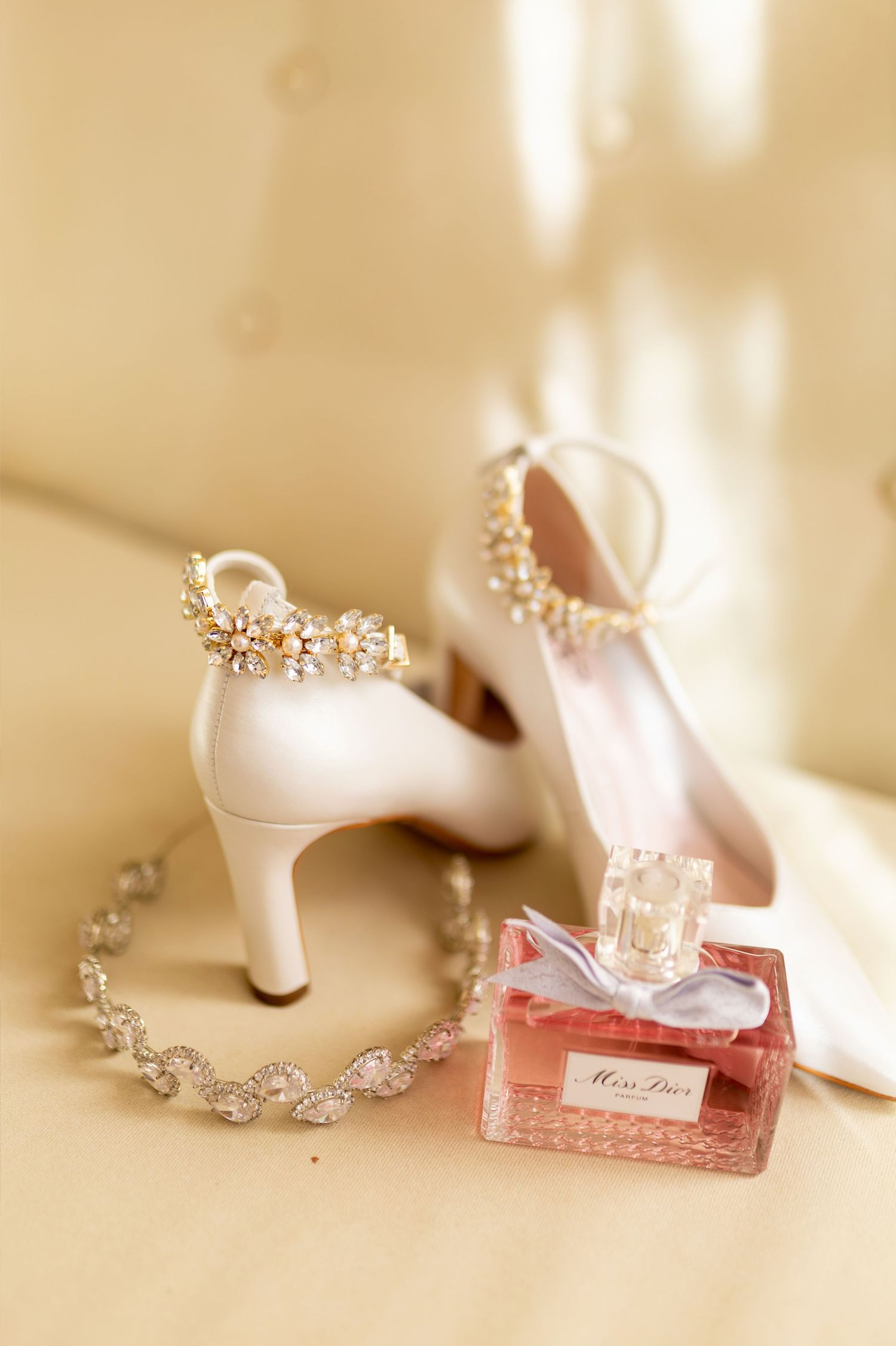 White bridal heels with crystal embellishments beside Miss Dior perfume and diamond bracelet on golden satin.