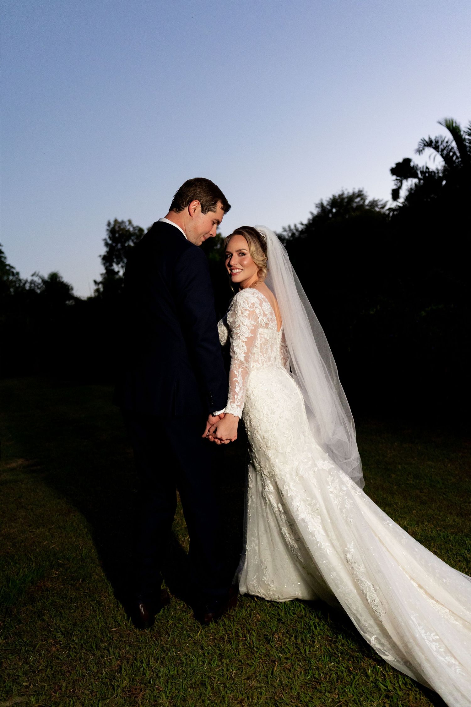 Bride in lace gown and veil holds hands with groom in dark suit outdoors at dusk.