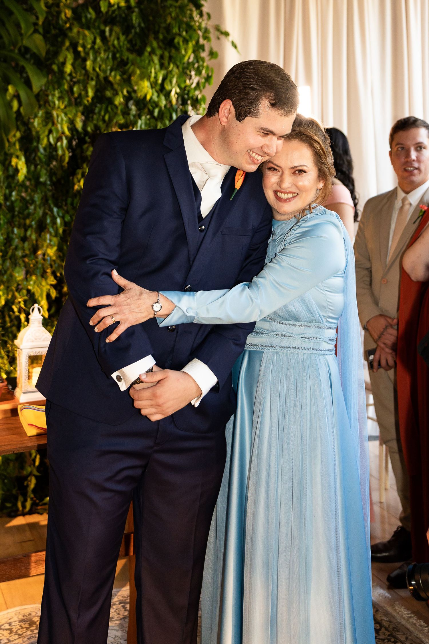 Couple embracing at a formal event, man in navy suit and woman in light blue gown, greenery backdrop.