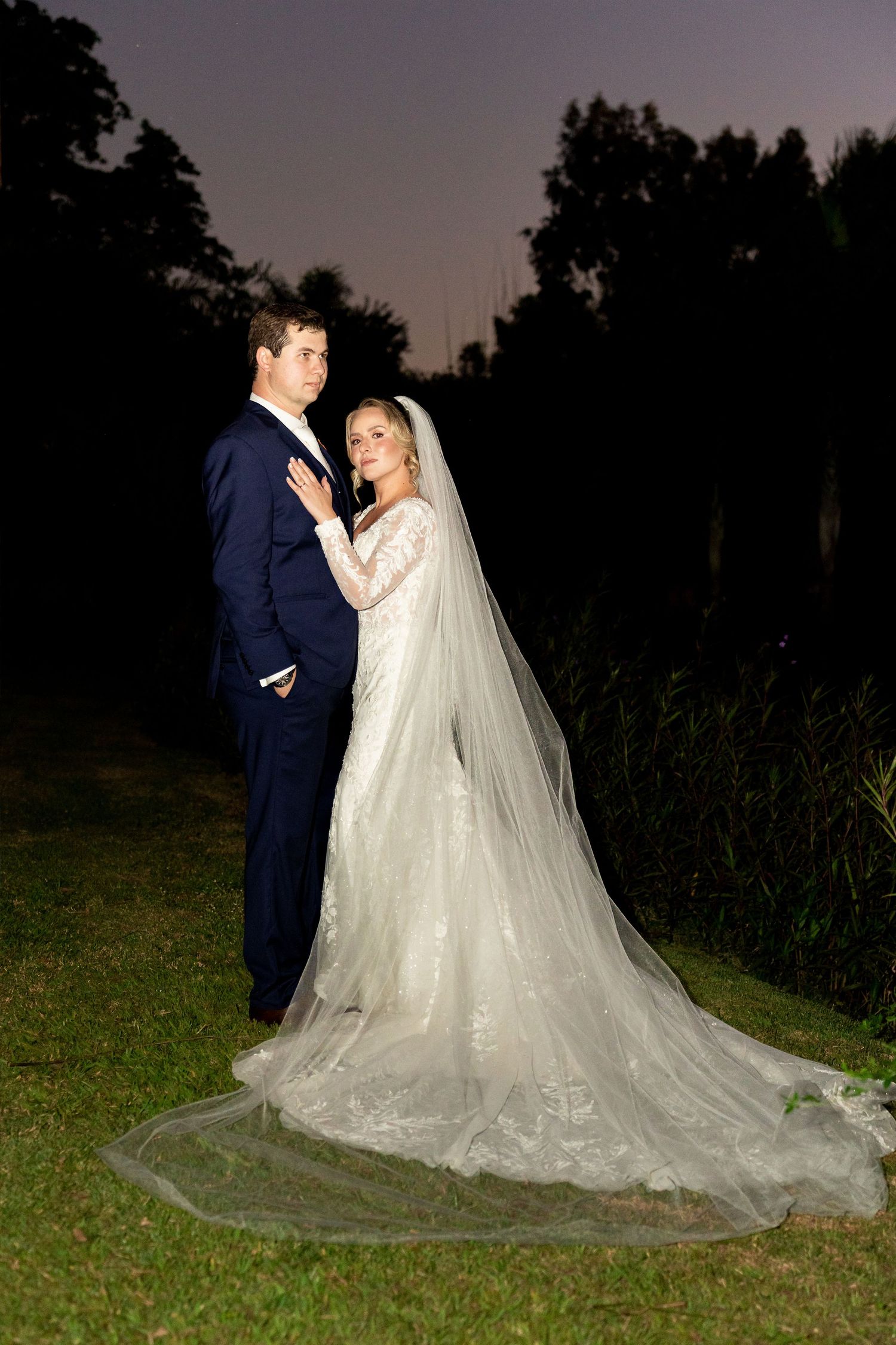 Bride in lace gown with cathedral veil and groom in navy suit pose outdoors at dusk.