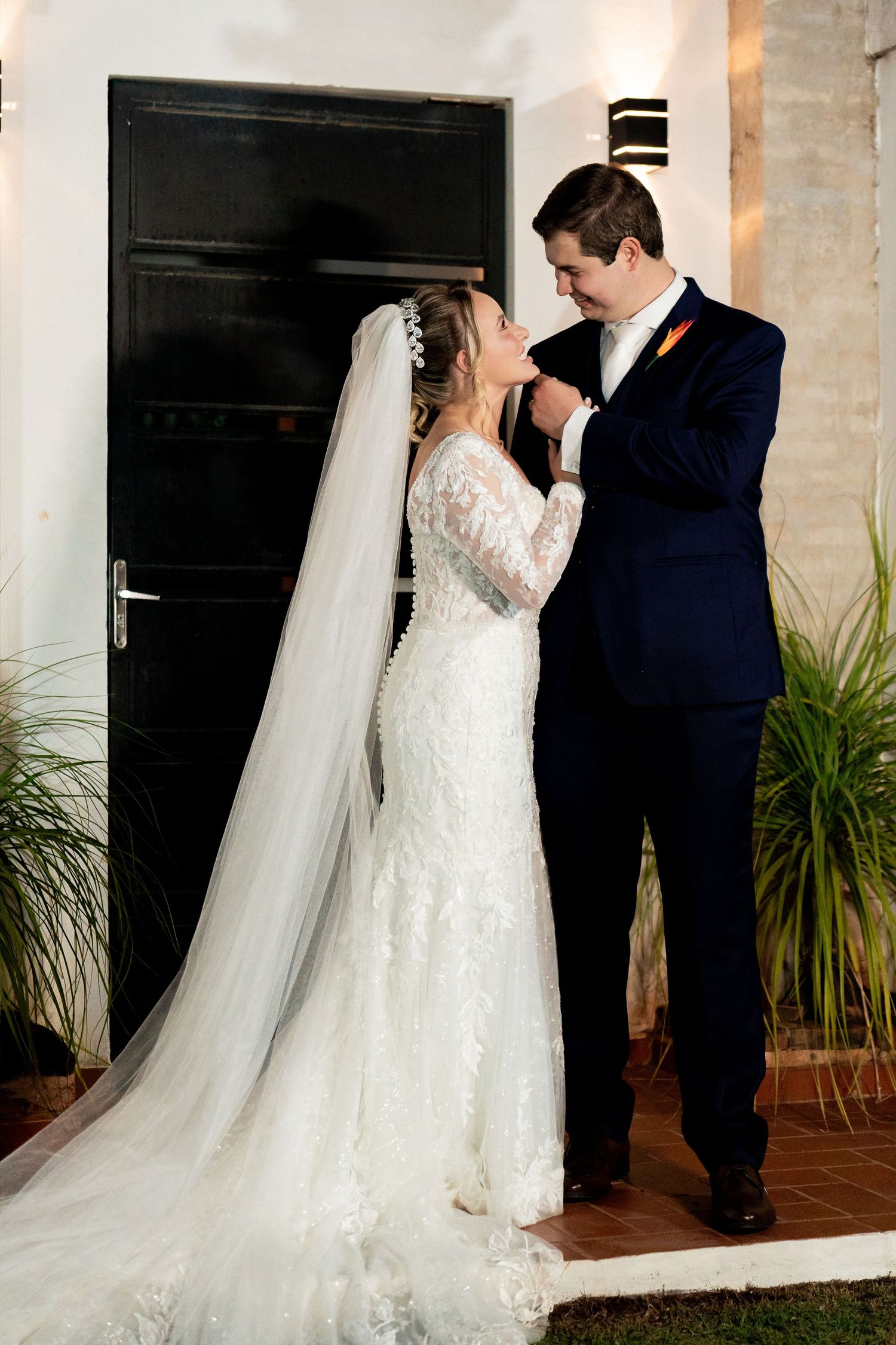 Groom in navy suit kisses bride's hand as she stands in lace wedding gown with long veil outside elegant venue.