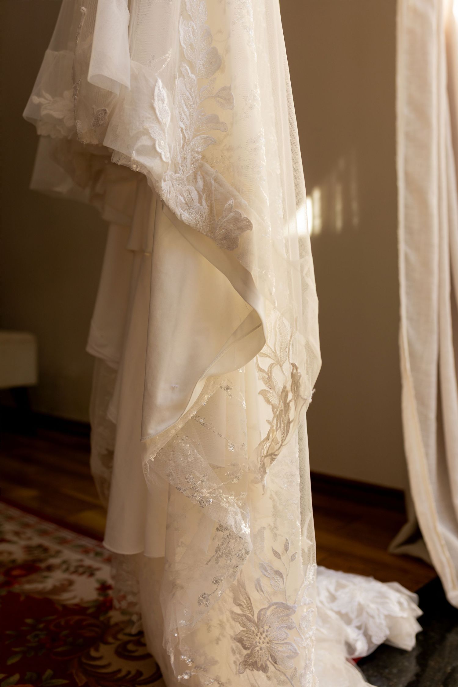 Elegant white lace wedding dress hanging, featuring floral embroidery and layered chiffon skirt in a softly lit room.