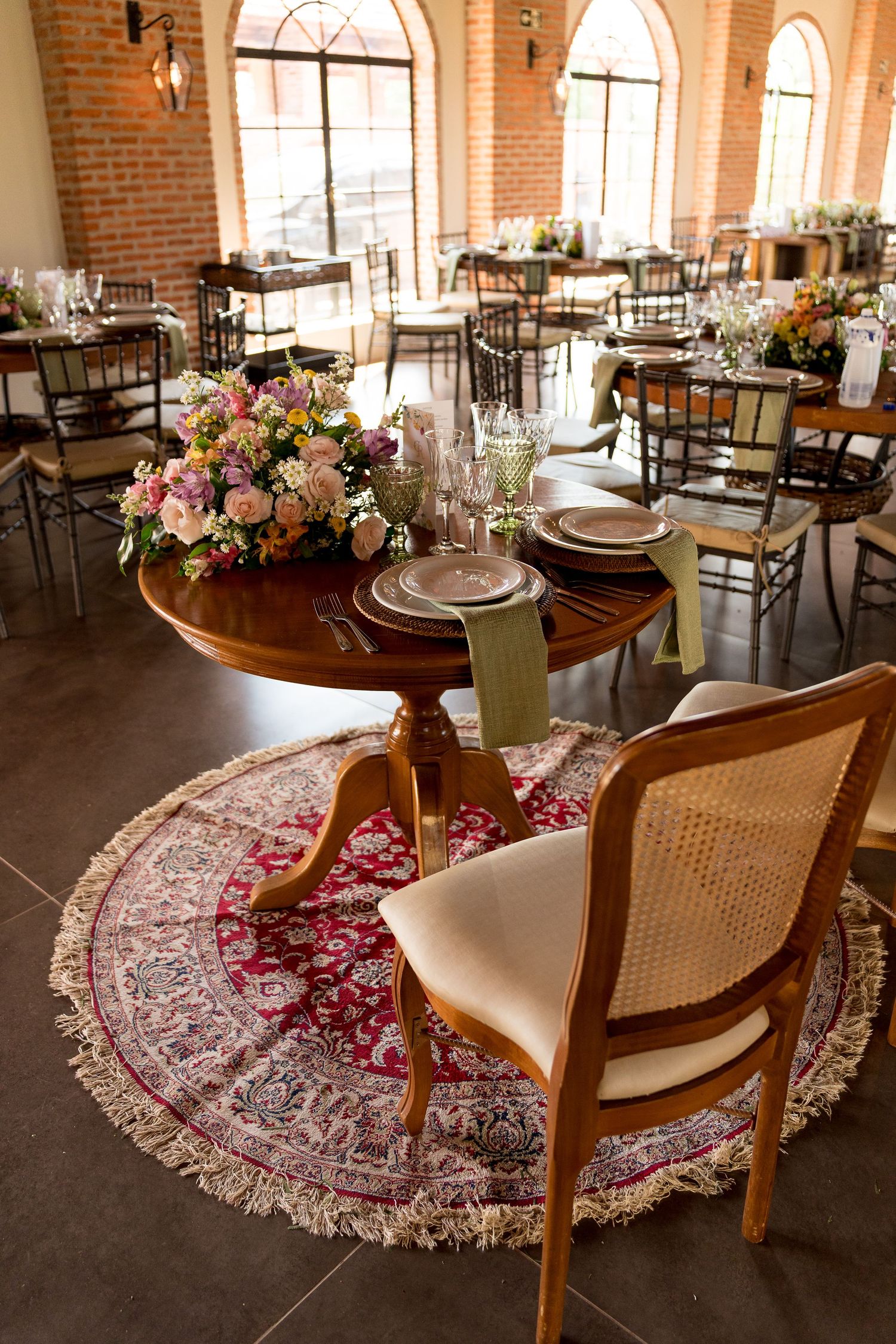 Elegant wedding reception venue with round wooden tables, floral centerpieces, chiavari chairs, and arched windows in a brick-walled room.