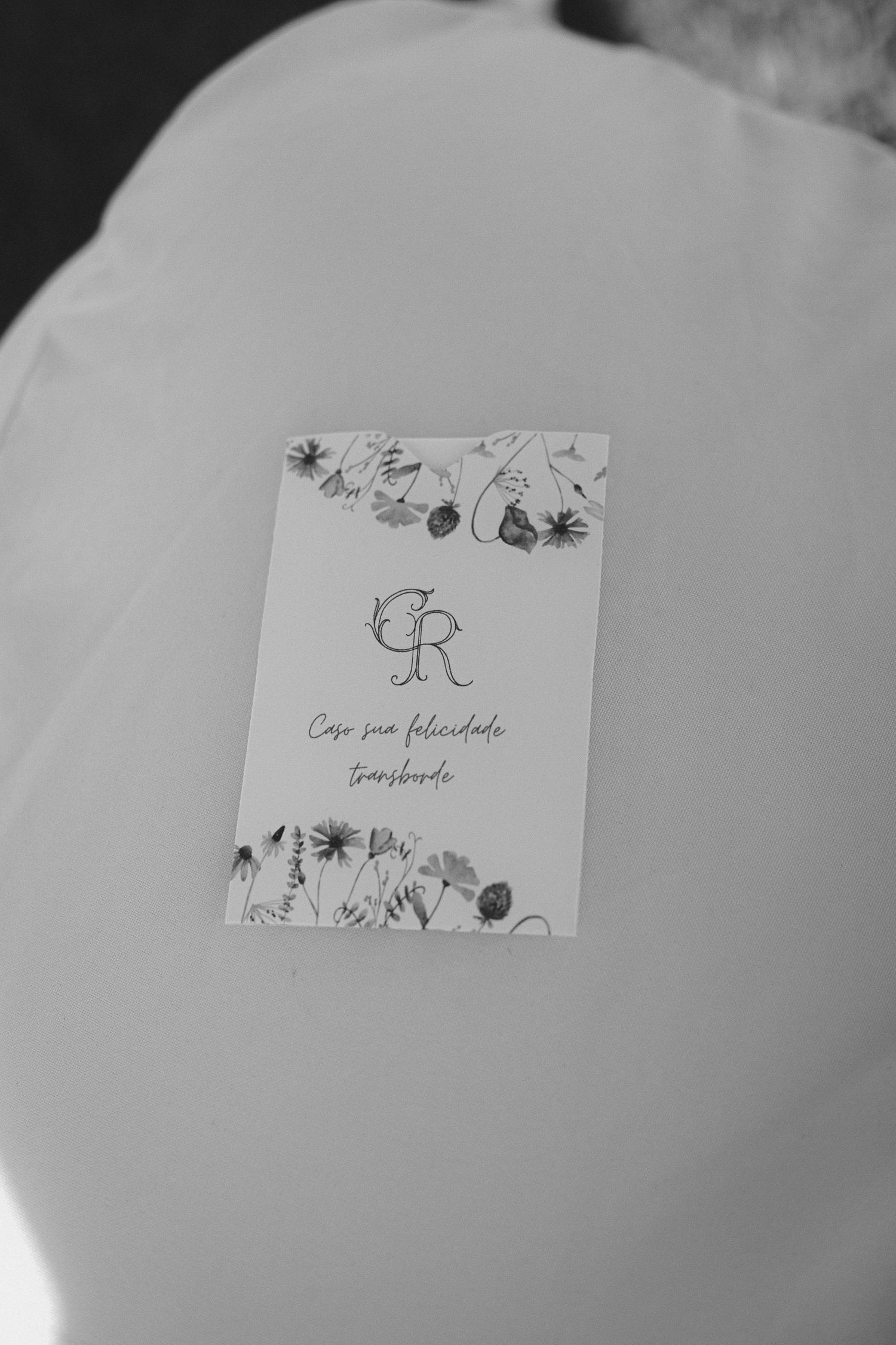 Black and white photo of a floral wedding card with monogram CR and Portuguese text on white fabric.