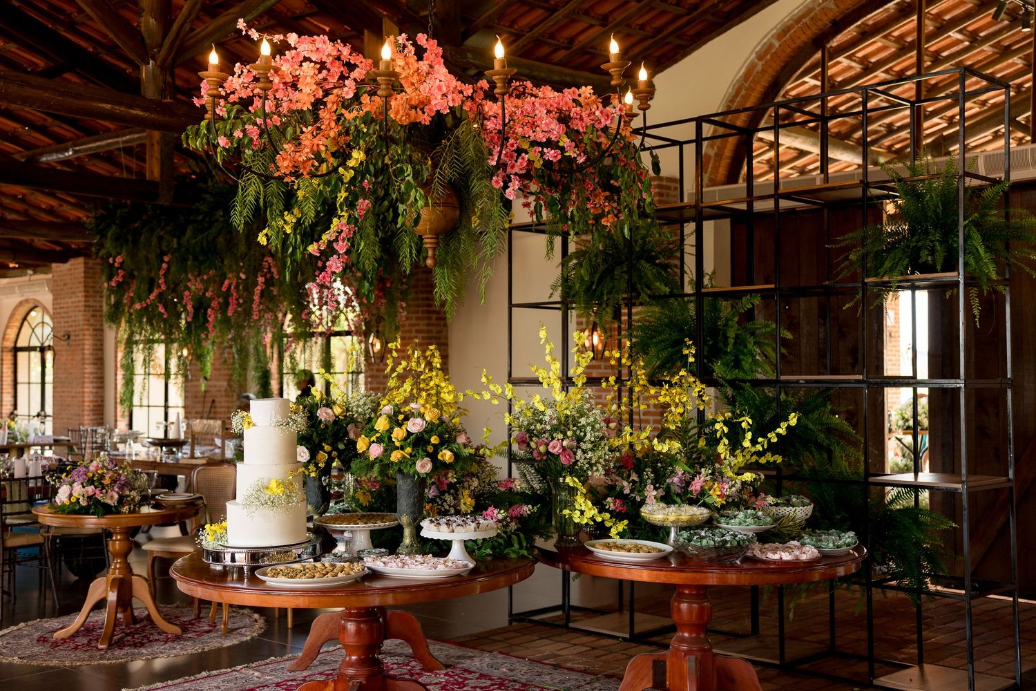 Elegant wedding dessert table adorned with lush floral arrangements, hanging pink blooms, and a tiered white cake in a rustic venue.