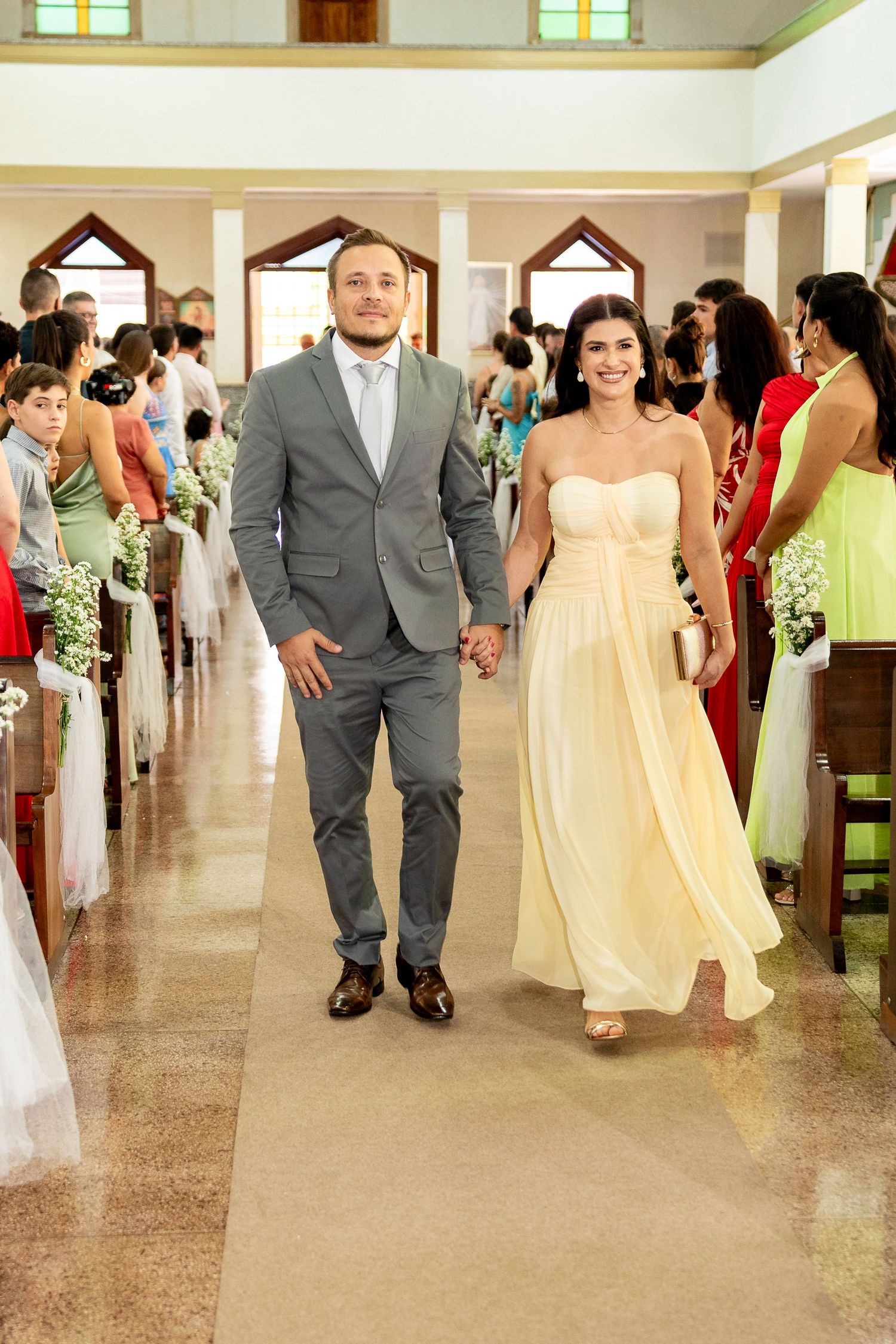 Couple walks down church aisle at wedding, man in gray suit, woman in yellow gown, guests seated in pews.