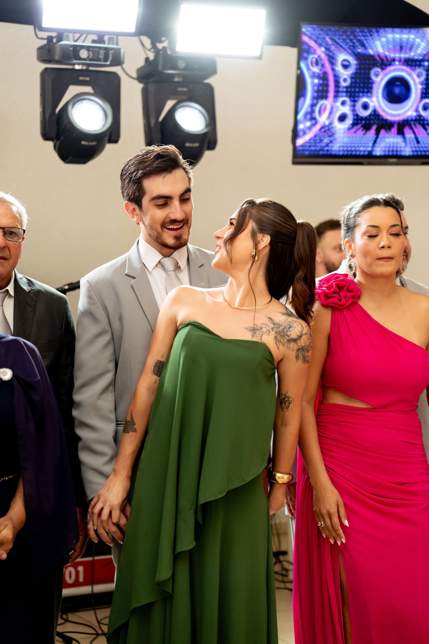 Couple in elegant attire, man in gray suit, woman in green gown with tattoos, beside woman in pink dress at event.